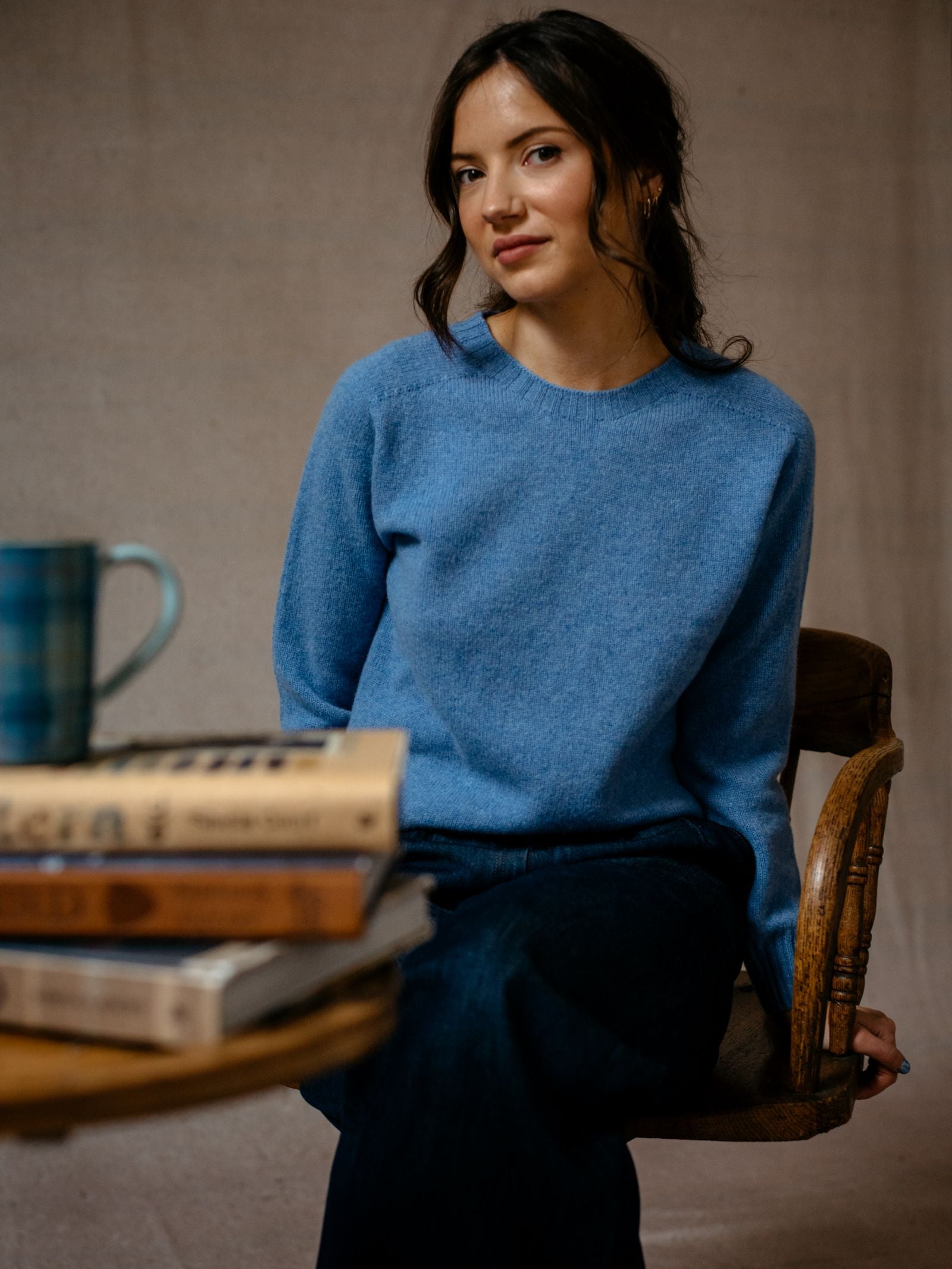 A woman in a blue Campbells of Beauly Geelong Lambswool Crew Neck Jumper, made in Scotland, sits on a wooden chair facing the camera. Stacked books and a blue plaid mug are on the table; the softly lit background is neutral.