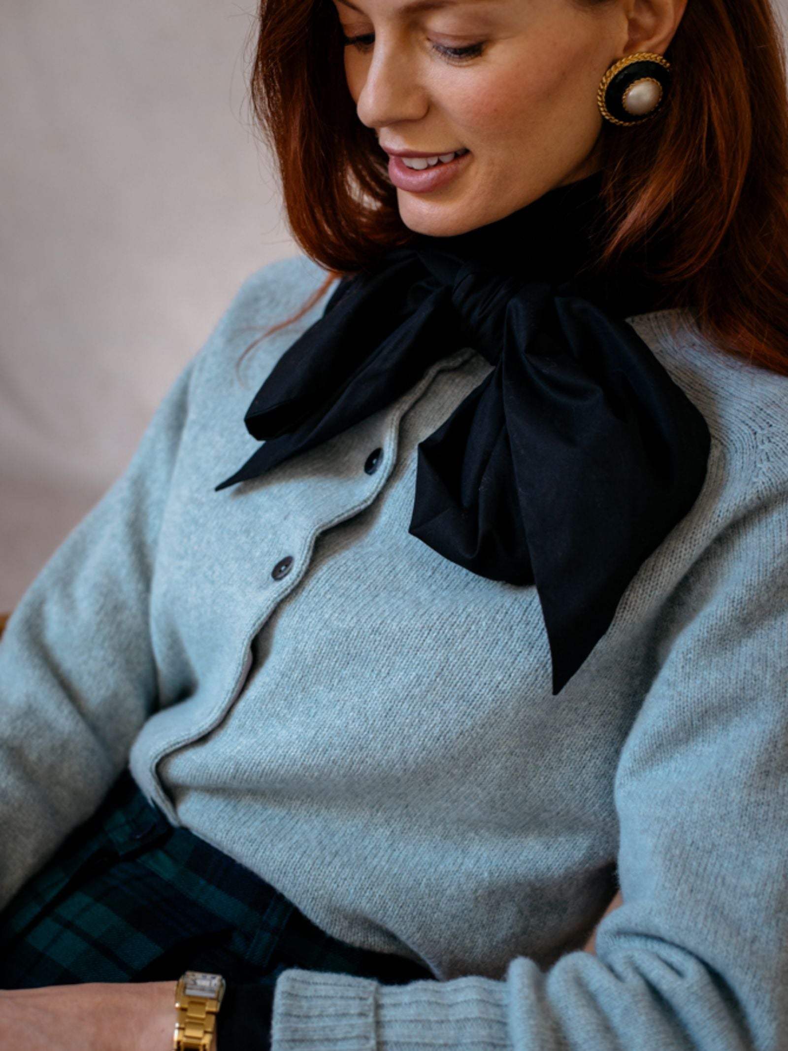 A woman with long auburn hair smiles softly and looks down, wearing a light blue Campbells of Beauly Crew Cardigan, a large black bow scarf, pearl earrings with gold trim, and a gold wristwatch.