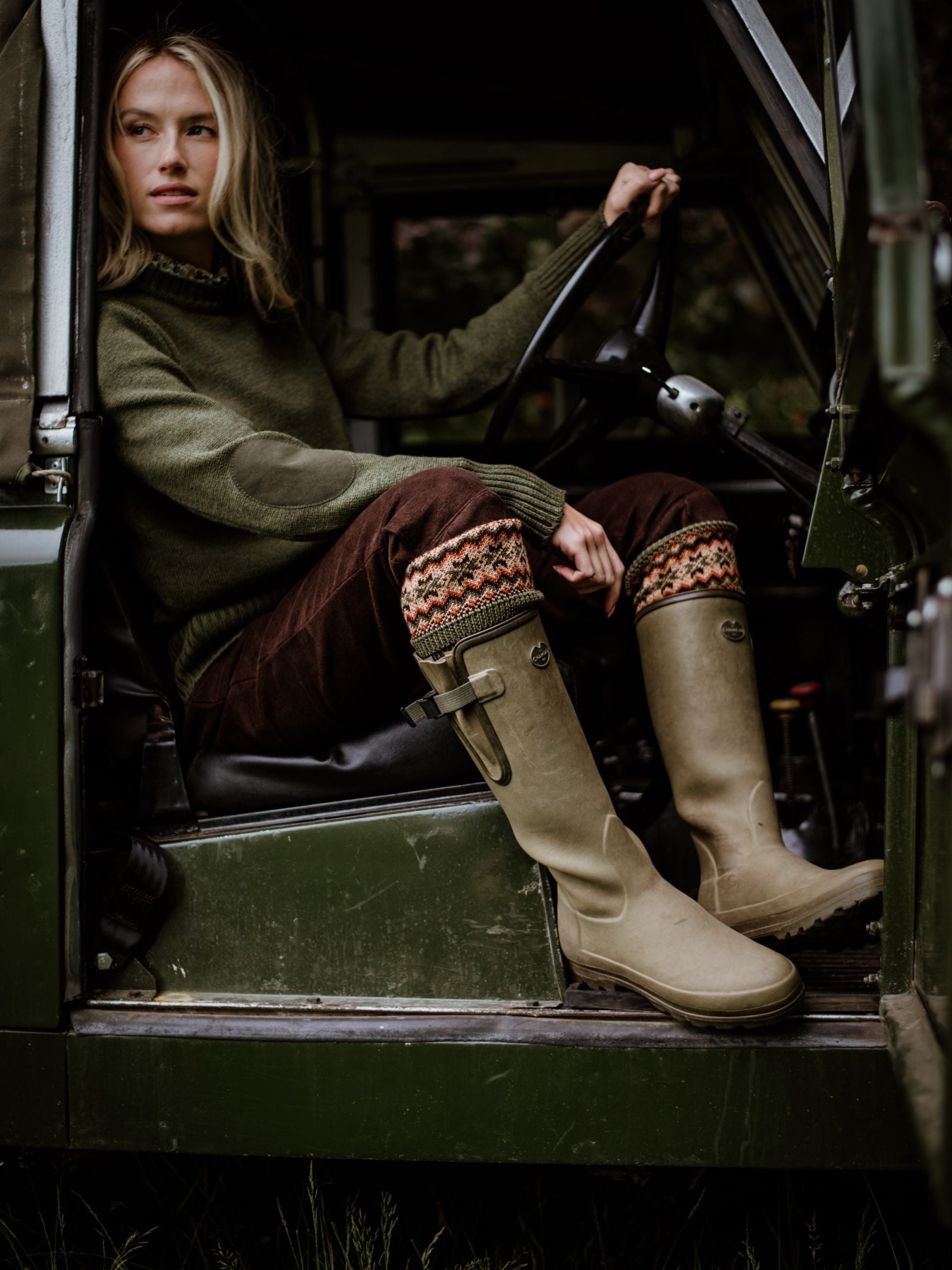 A woman in an olive green sweater, Campbells of Beauly Merino Fairisle Shooting Socks, and beige boots sits in the driver’s seat of a green vehicle, holding the steering wheel and gazing sideways.