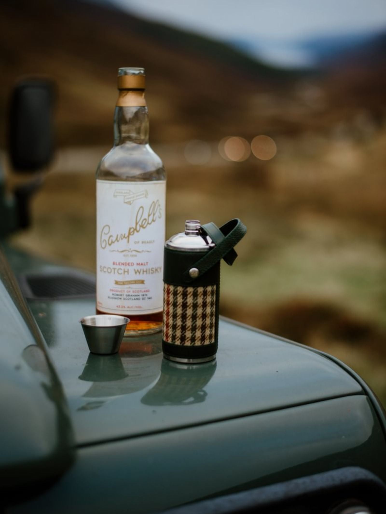 An 8oz Forest Green Hunter Flask by Campbell's of Beauly x Ettinger, a small metal cup, and a bottle of Scotch whisky rest on a green vehicle’s hood, highlighting British craftsmanship against a blurred mountain backdrop.