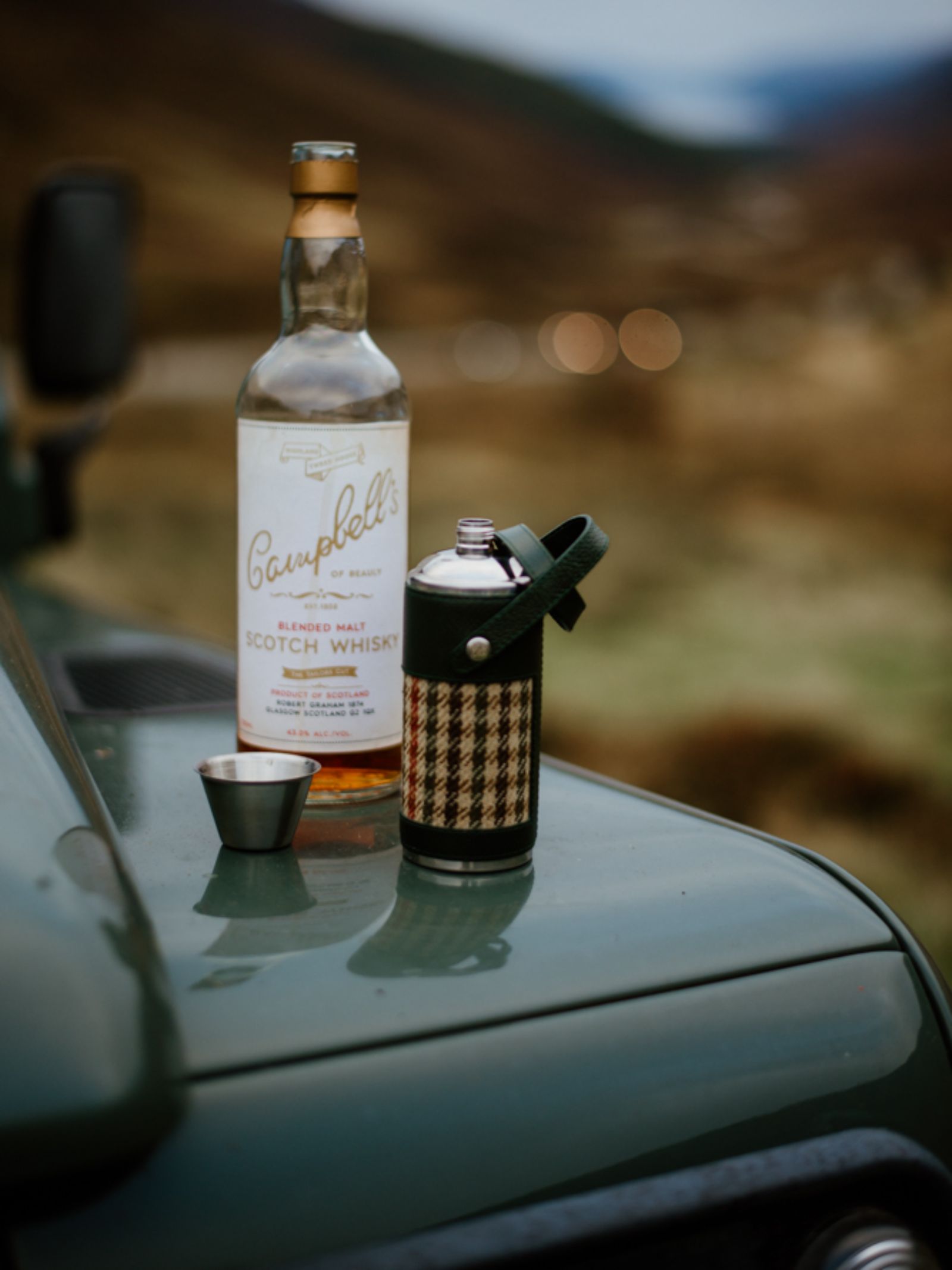 A bottle of Campbell’s Scotch whisky, a metal cup, and the 8oz Bordeaux Hunter Flask by Campbell's of Beauly x Ettinger—known for exceptional British craftsmanship—rest on the hood of a green vehicle with a blurred natural background.