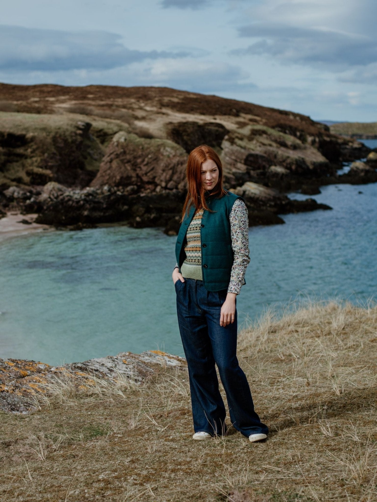 A woman with long red hair stands on grassy terrain by cliffs and blue water, wearing a patterned blouse, blue trousers, and the Campbells of Beauly Quilted Buttoned Gilet, gazing to the side under a cloudy sky.