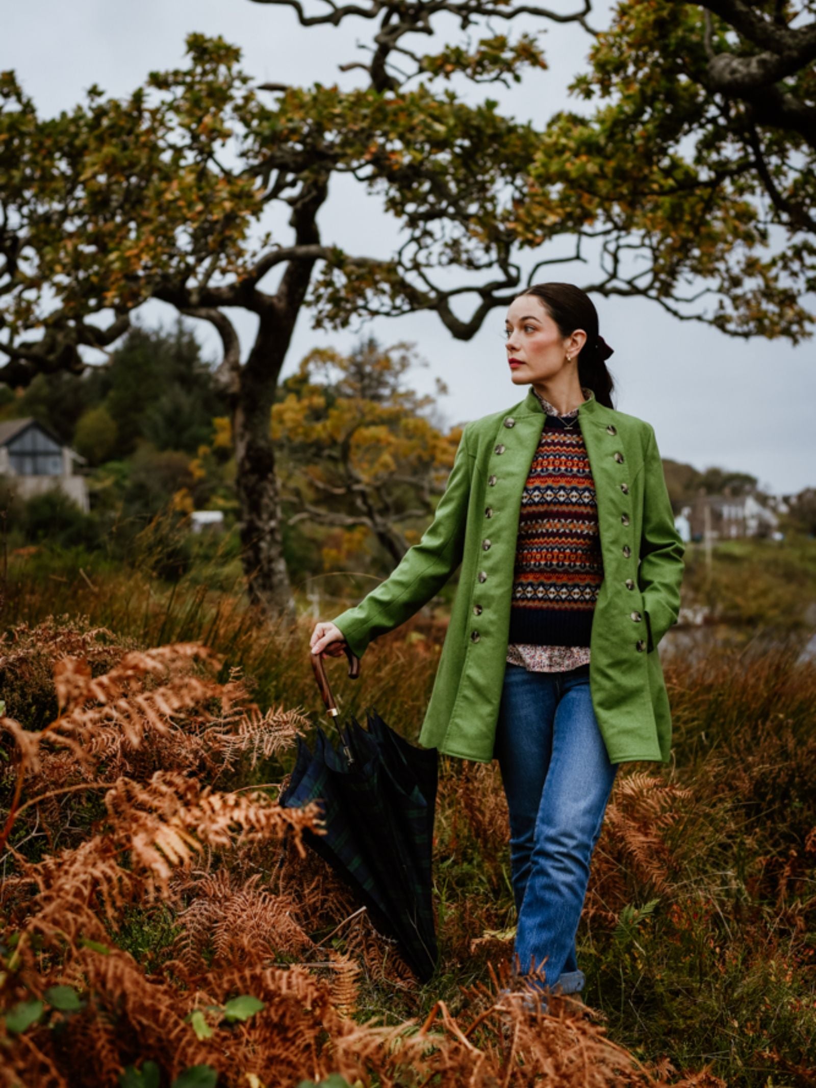 A woman in a green Campbell's of Beauly Pirate Coat and blue jeans stands outdoors among brown ferns, holding a closed umbrella. She looks to the side with trees and houses visible in the cloudy background.