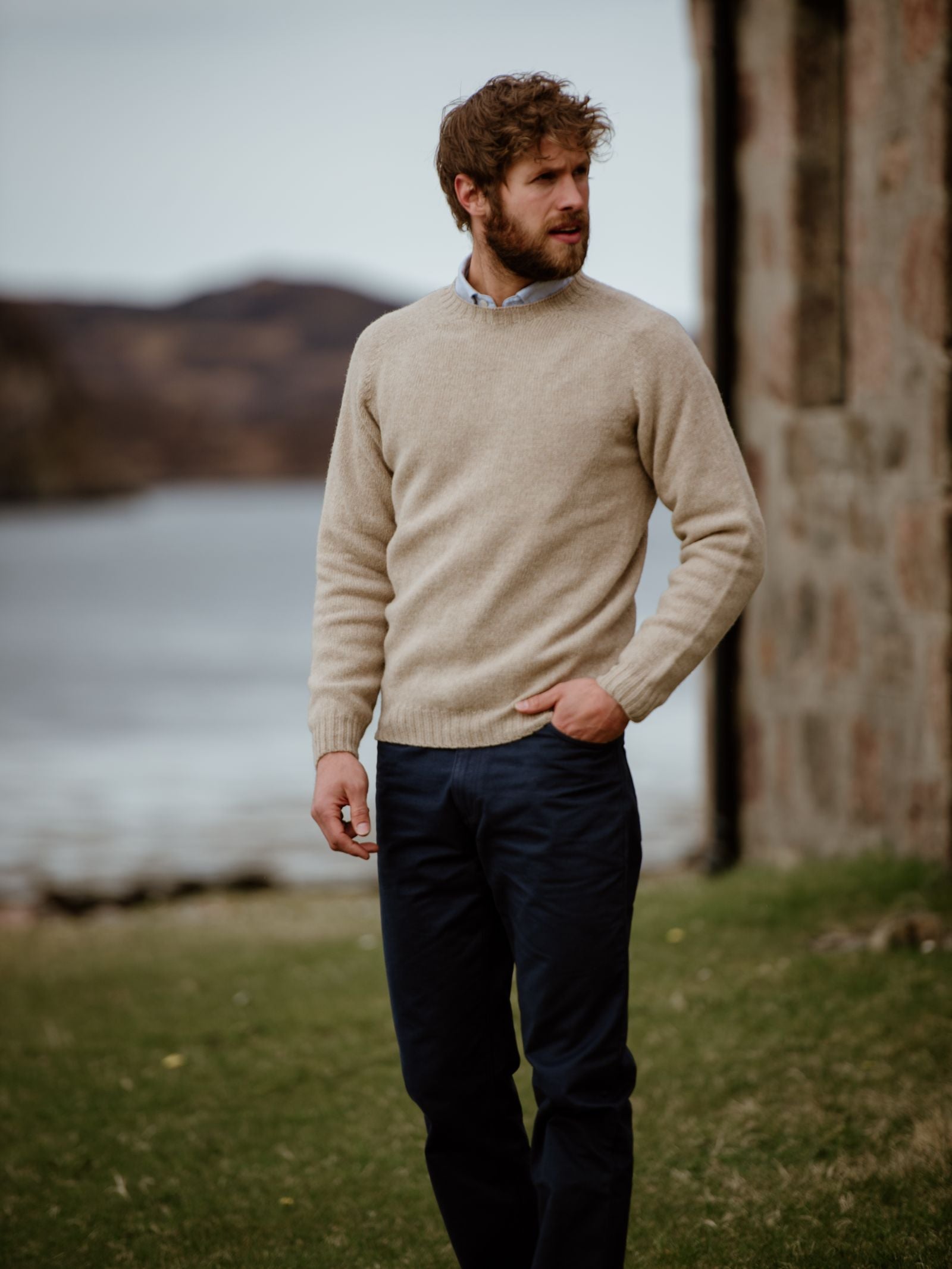 A bearded man in dark trousers wears a beige Campbells of Beauly Shetland Jumper, crafted from soft lambswool yarns, standing outdoors near a stone building with water and hills in the background.