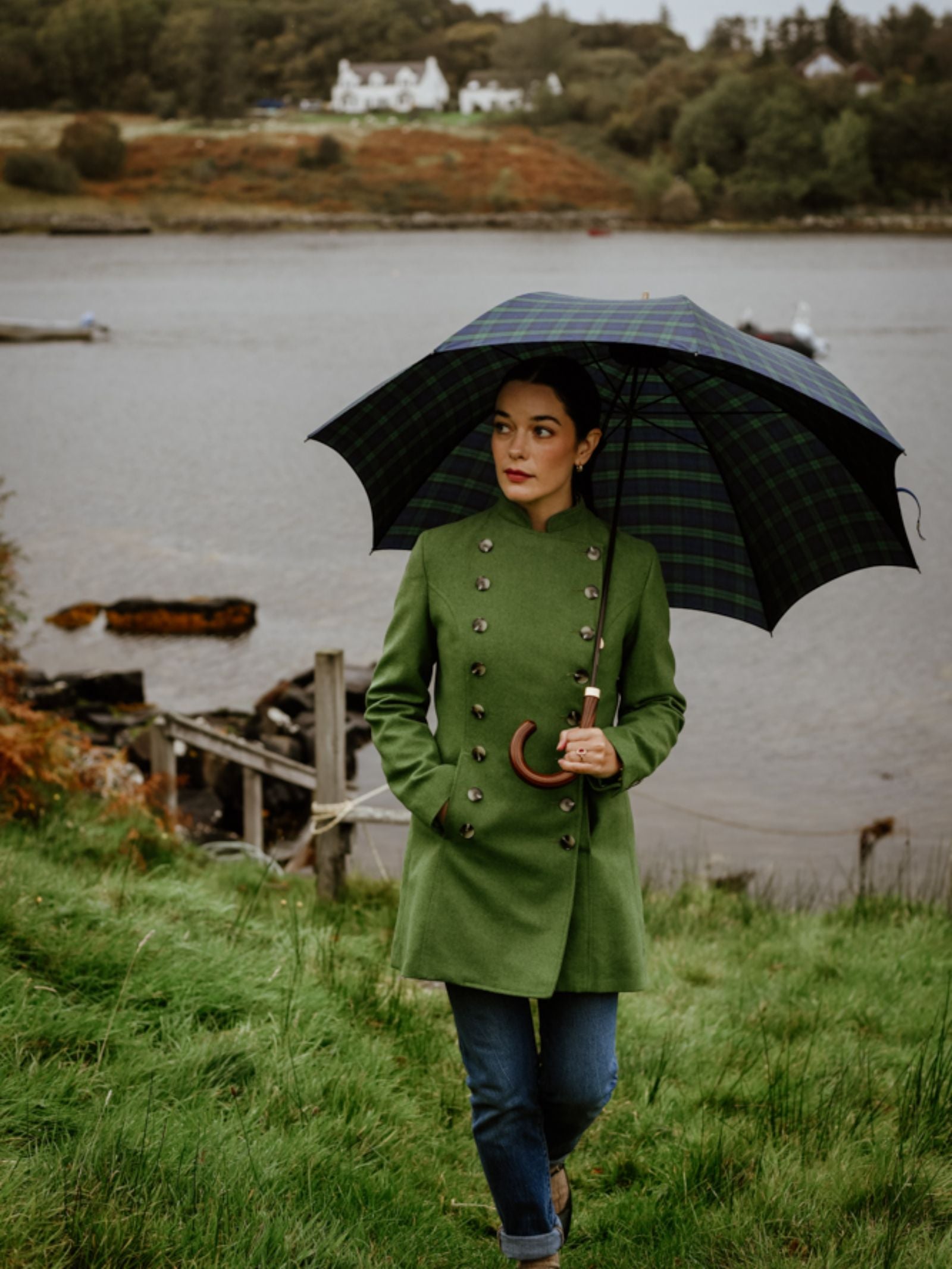 A woman in the Campbell's of Beauly Pirate Coat and jeans holds a plaid umbrella while walking on grass near a lake, with trees and houses in the background under an overcast sky.