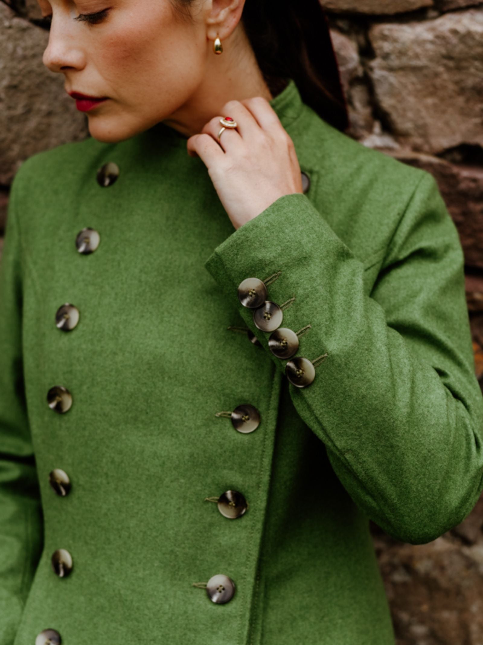 A woman in a tailored green Campbell's of Beauly Pirate Coat with large decorative buttons stands by a stone wall, her hand near her collar, showing a gold earring and red lipstick—outerwear that makes a true statement.