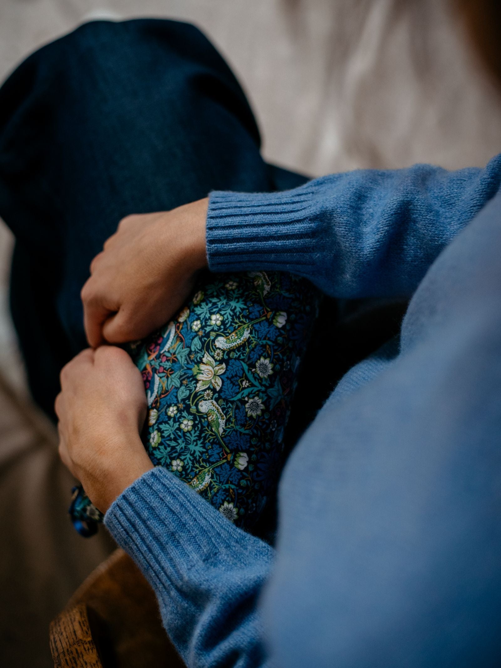 Wearing a Campbells of Beauly Geelong Lambswool Crew Neck Jumper and jeans, a person sits with clasped hands over a knee draped in blue floral fabric, creating a calm, introspective scene.