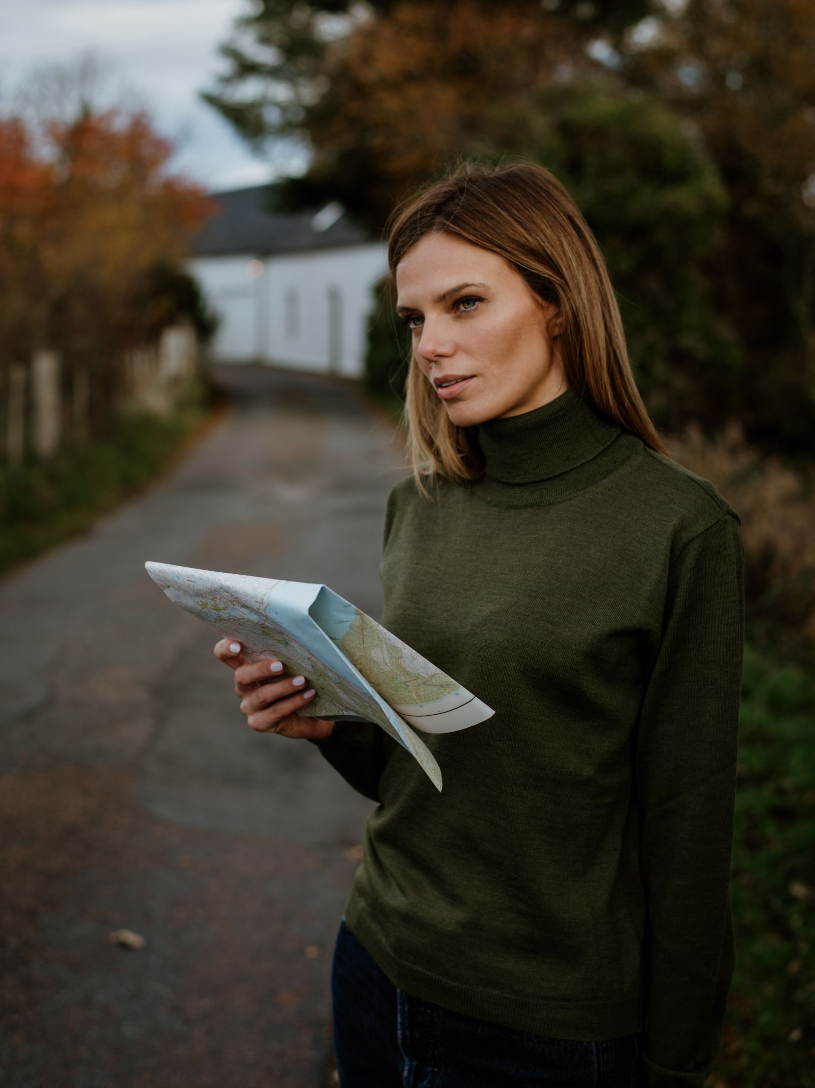 A woman wears the Campbells of Beauly Merino Poloneck in green, holding a folded map on a rural road with autumn trees and a white building behind her—thoughtfully styled for crisp fall days.