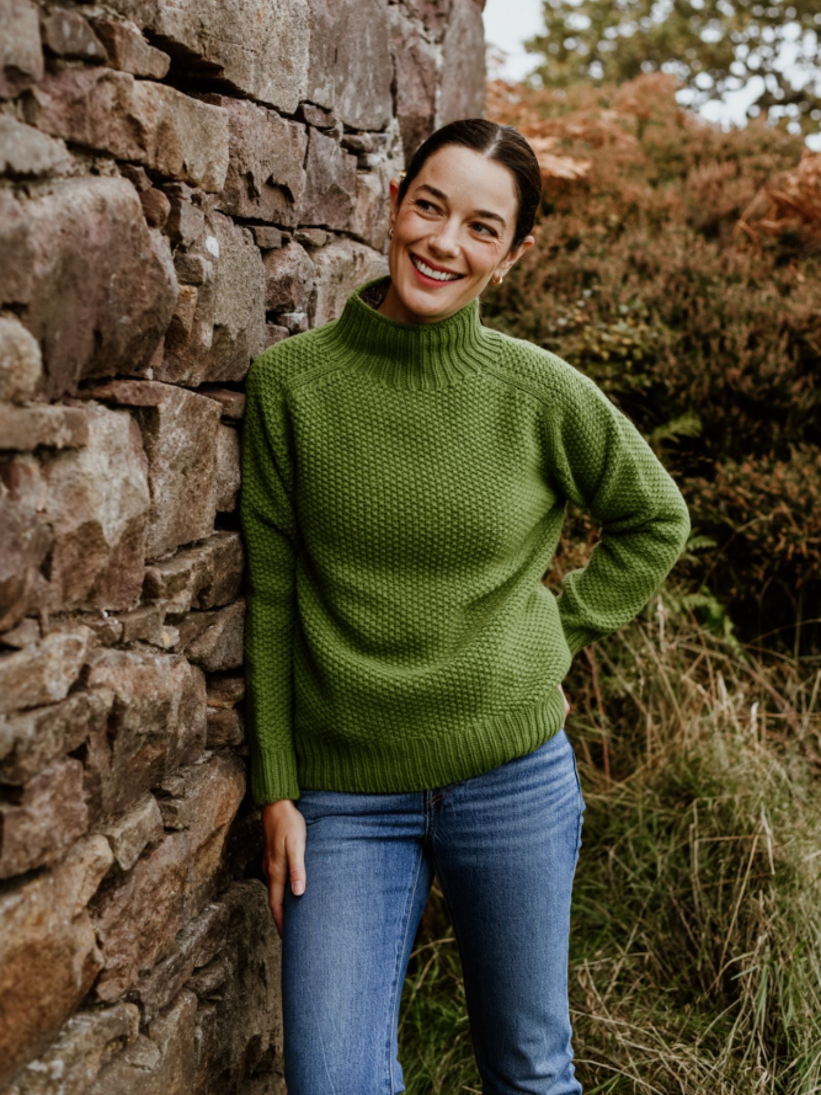 A woman smiles outdoors by a stone wall with one hand in her pocket, wearing the Campbell's of Beauly Chunky Moss Stitch Polo and blue jeans. Bushes and grass are visible in the background.