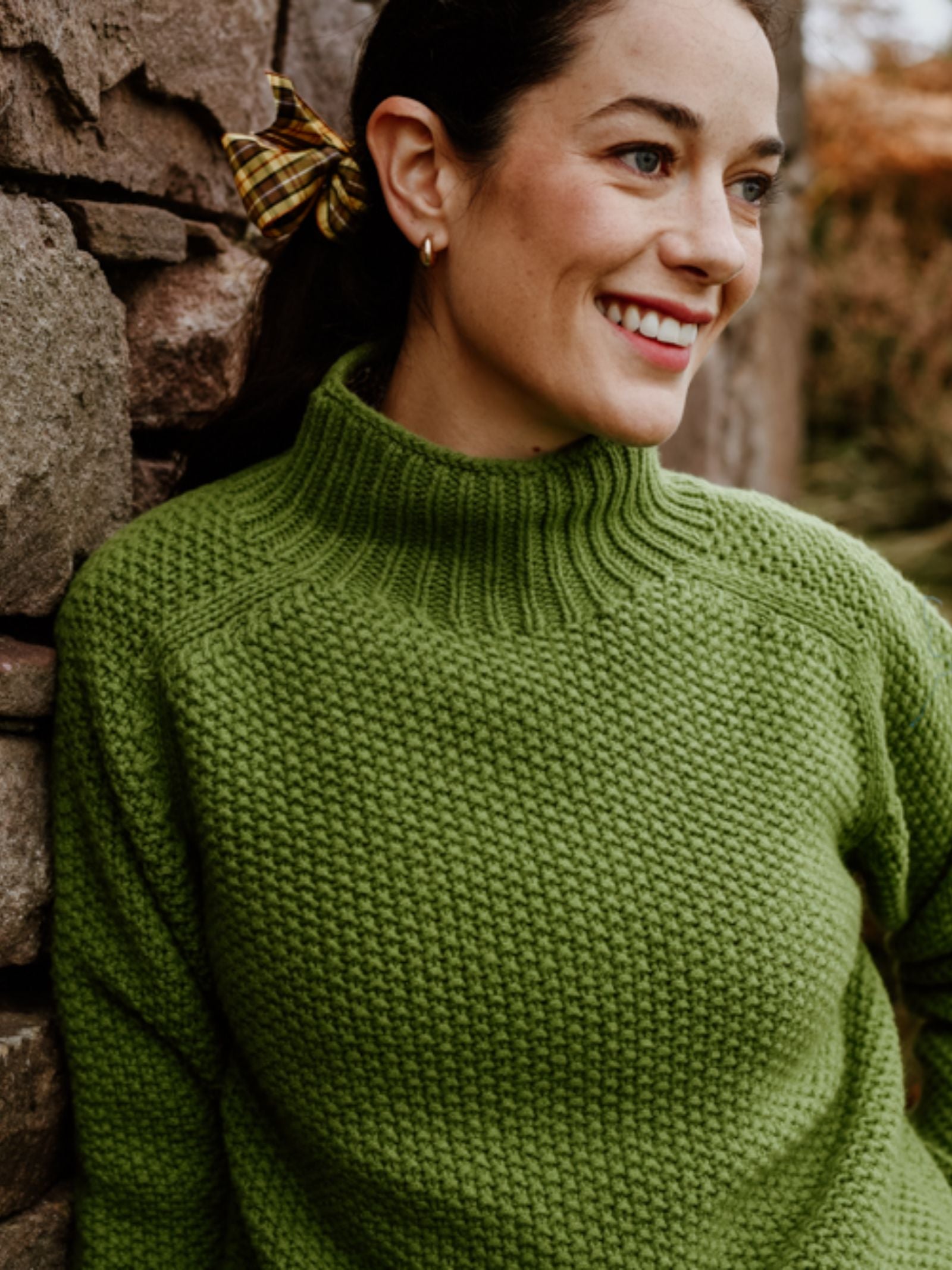 A woman with dark hair in a plaid hair tie and gold hoop earrings smiles as she leans against a stone wall outdoors, wearing the Chunky Moss Stitch Polo by Campbell's of Beauly.