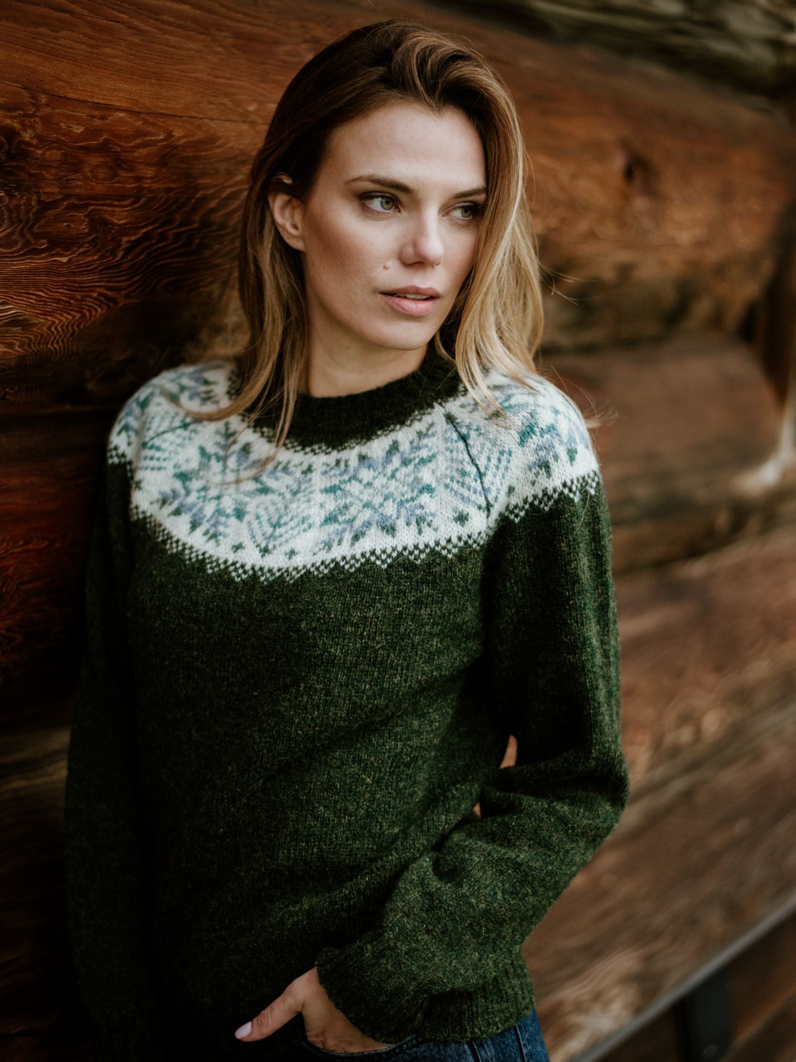 A woman with long, light brown hair wears the Campbells of Beauly Shetland Wool Snowflake Crew Jumper in dark green as she stands in front of a wooden wall, gazing thoughtfully to the side.