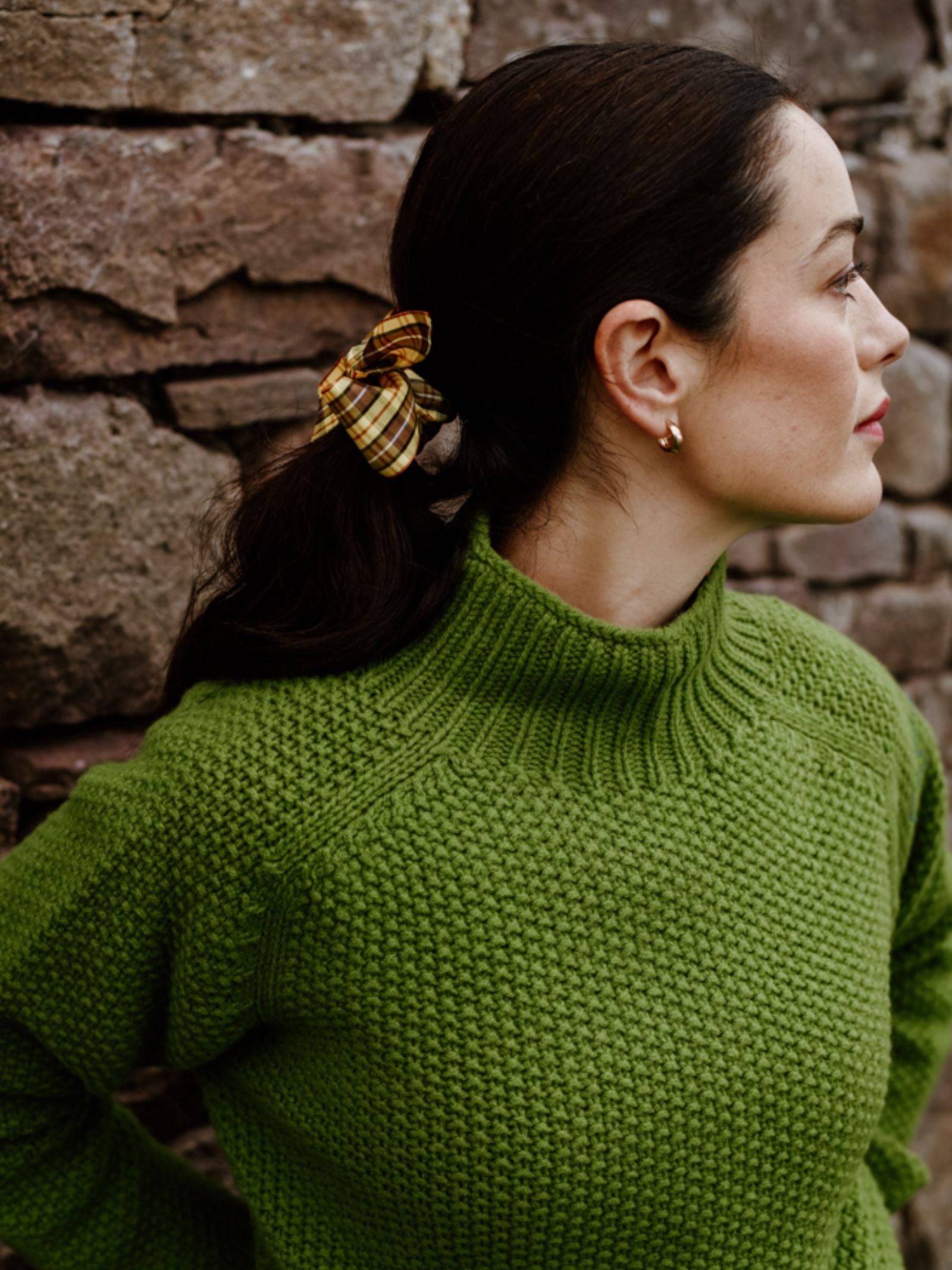 A woman with dark hair tied back with a yellow plaid scrunchie, wearing the Campbell's of Beauly Chunky Moss Stitch Polo in bright green, stands before a stone wall and looks to the right.