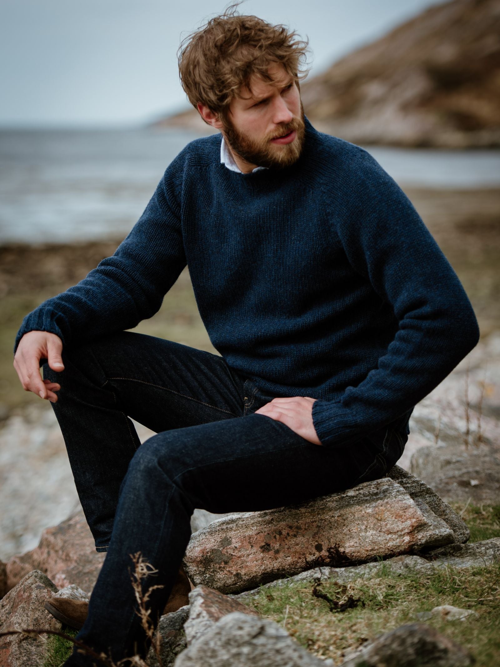 A man with brown hair and a beard, wearing a navy blue Campbells of Beauly Geelong Lambswool Chunky Crew Jumper and dark jeans, sits on coastal rocks with water and hills in the background, looking to the side.