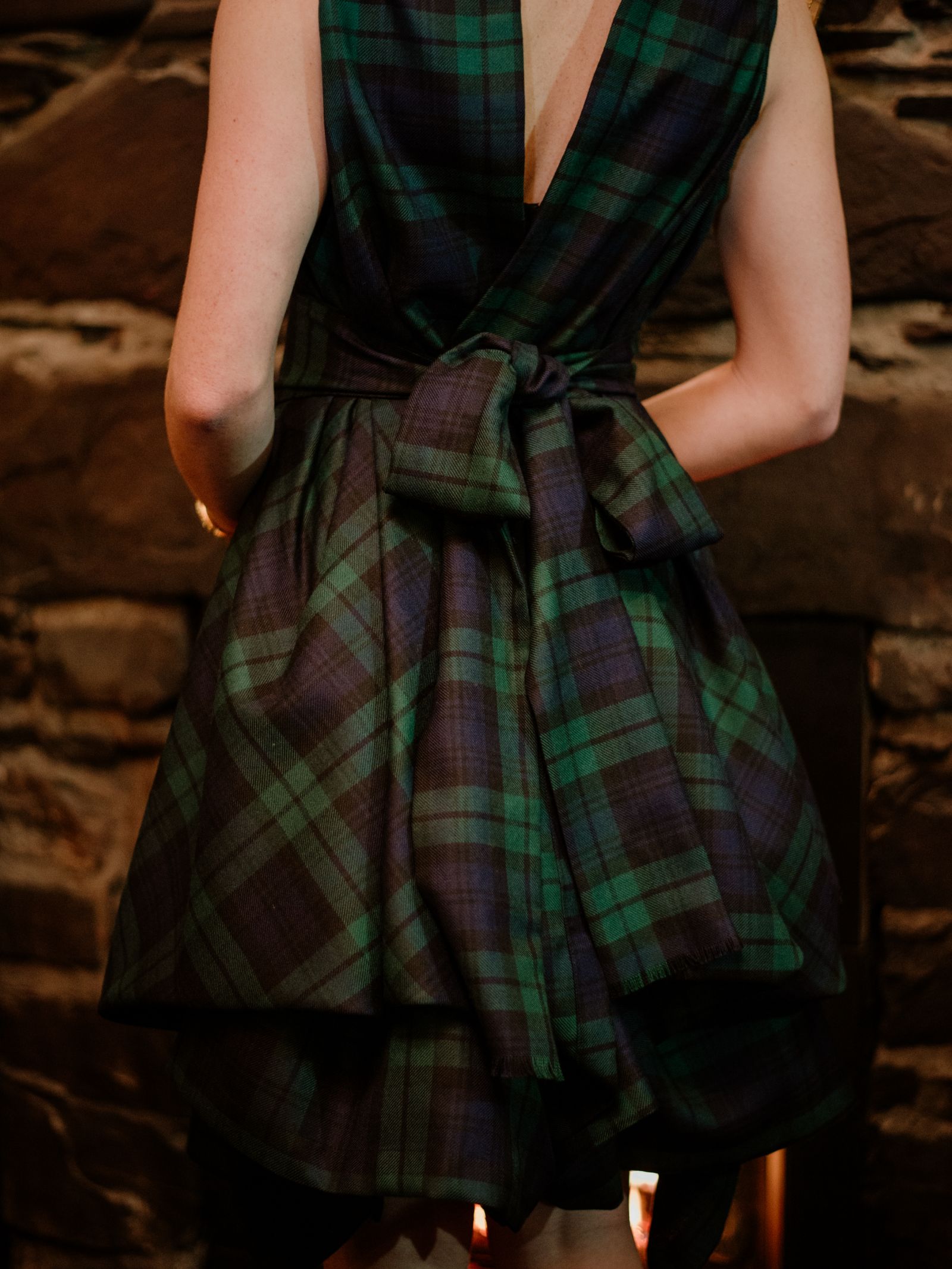 A person wears the Campbells of Beauly Reversible Wool Wrap Dress, green and navy plaid with a large back bow, inspired by Lochcarron of Scotland, standing in front of a stone wall facing away from the camera.