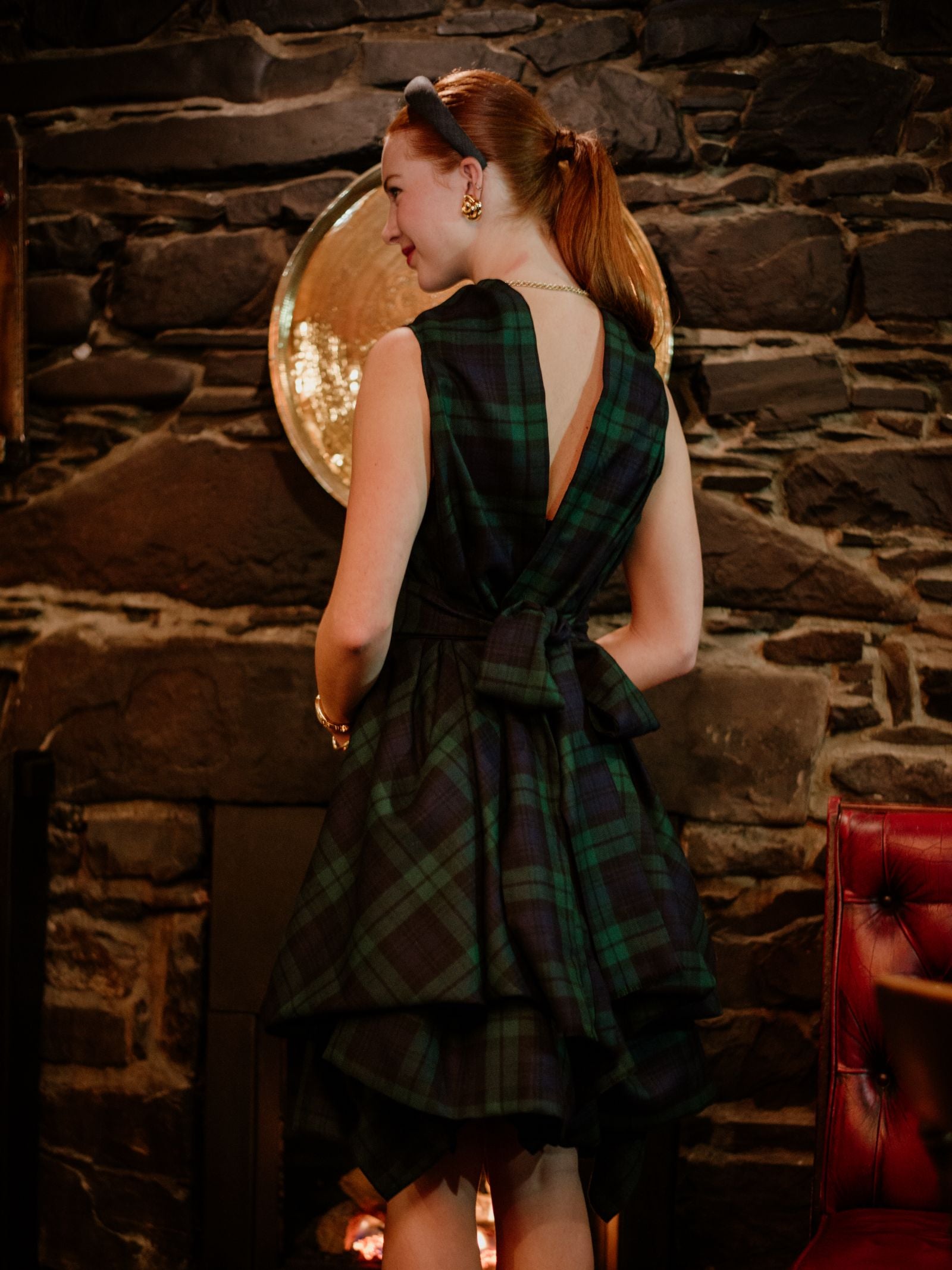 A woman with red hair in a ponytail stands with her back to the camera, wearing a Campbells of Beauly Reversible Wool Wrap Dress with a waist bow, in front of a stone wall and fireplace.