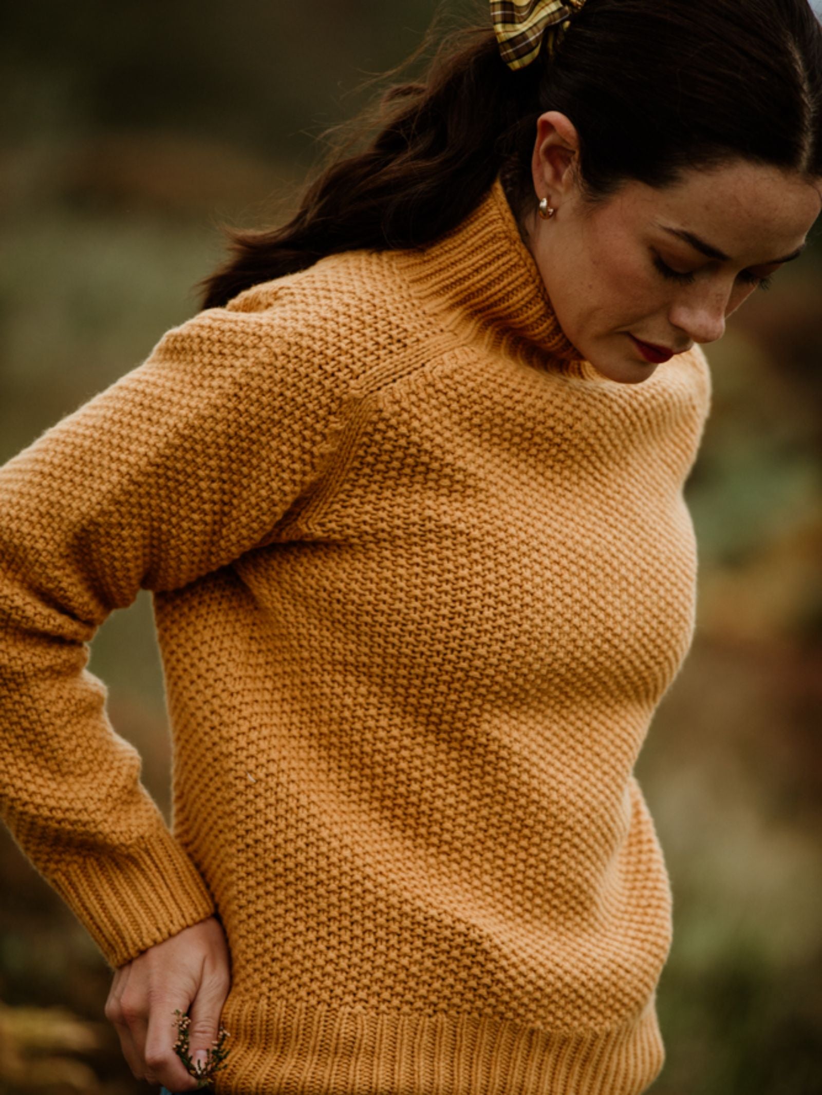 A woman with dark hair tied back by a ribbon looks down while wearing the Chunky Moss Stitch Polo from Campbell’s of Beauly, a mustard yellow chunky-knit turtleneck sweater. The blurred background suggests an outdoor setting.