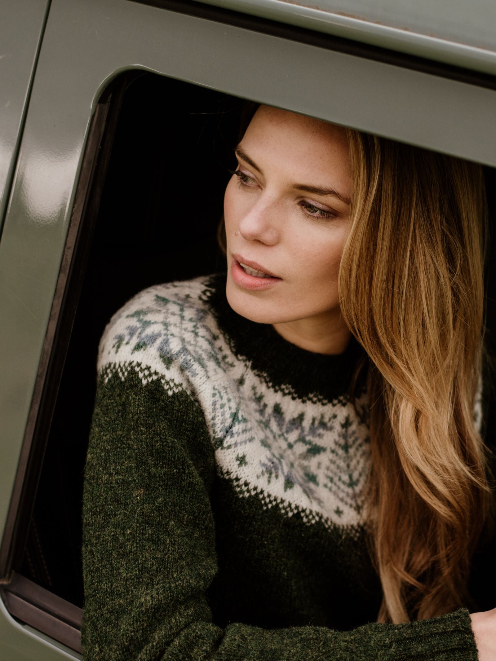 A woman with long, light brown hair, wearing the Campbells of Beauly Shetland Wool Snowflake Crew Jumper in dark green with a white and blue Fairisle pattern, looks thoughtfully out the open window of a green vehicle.