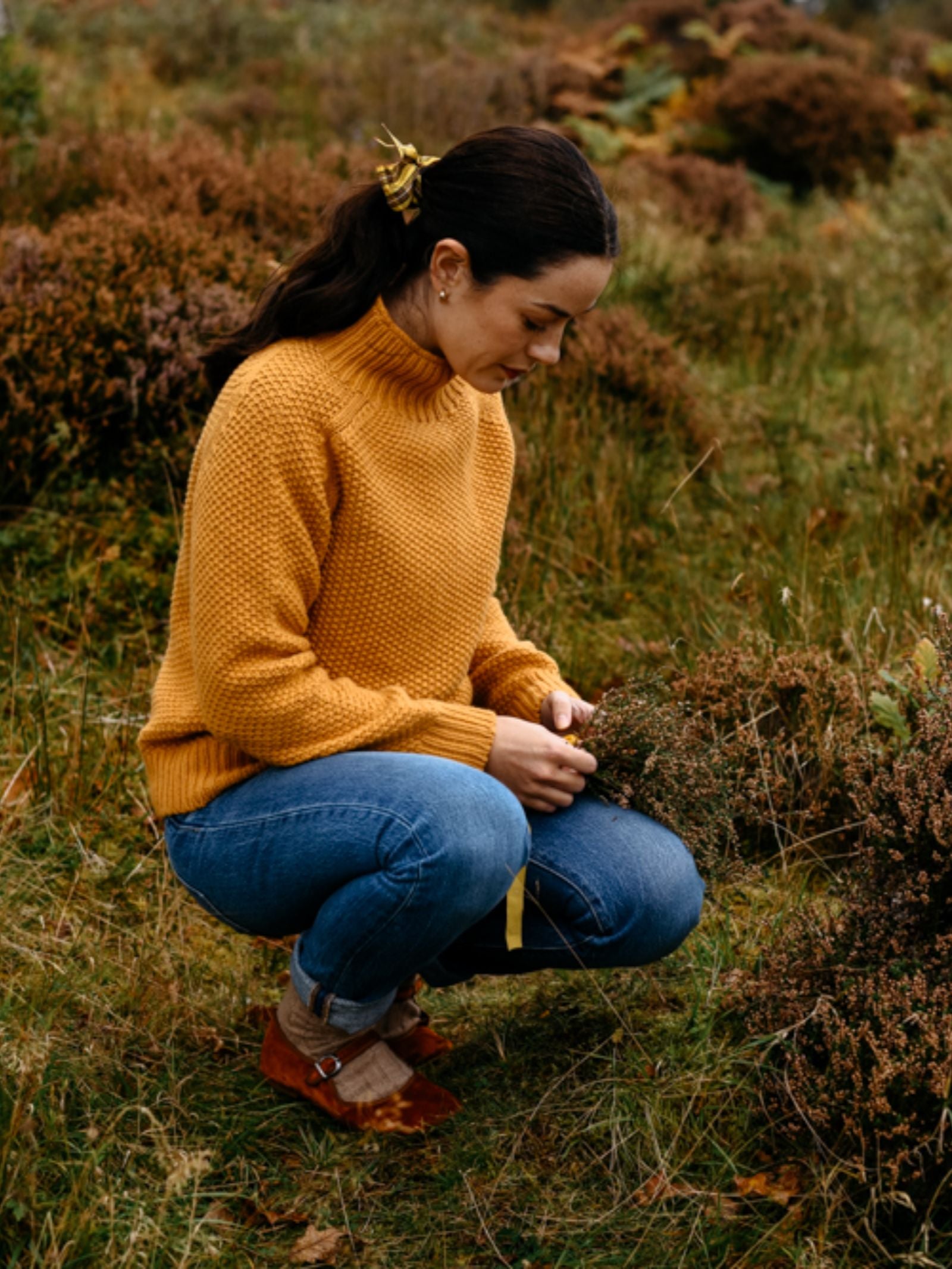 A woman wears the Chunky Moss Stitch Polo by Campbell's of Beauly with blue jeans as she crouches in a grassy field, gathering plants. Her dark hair is tied back with a yellow accessory as she focuses intently on the greenery before her.