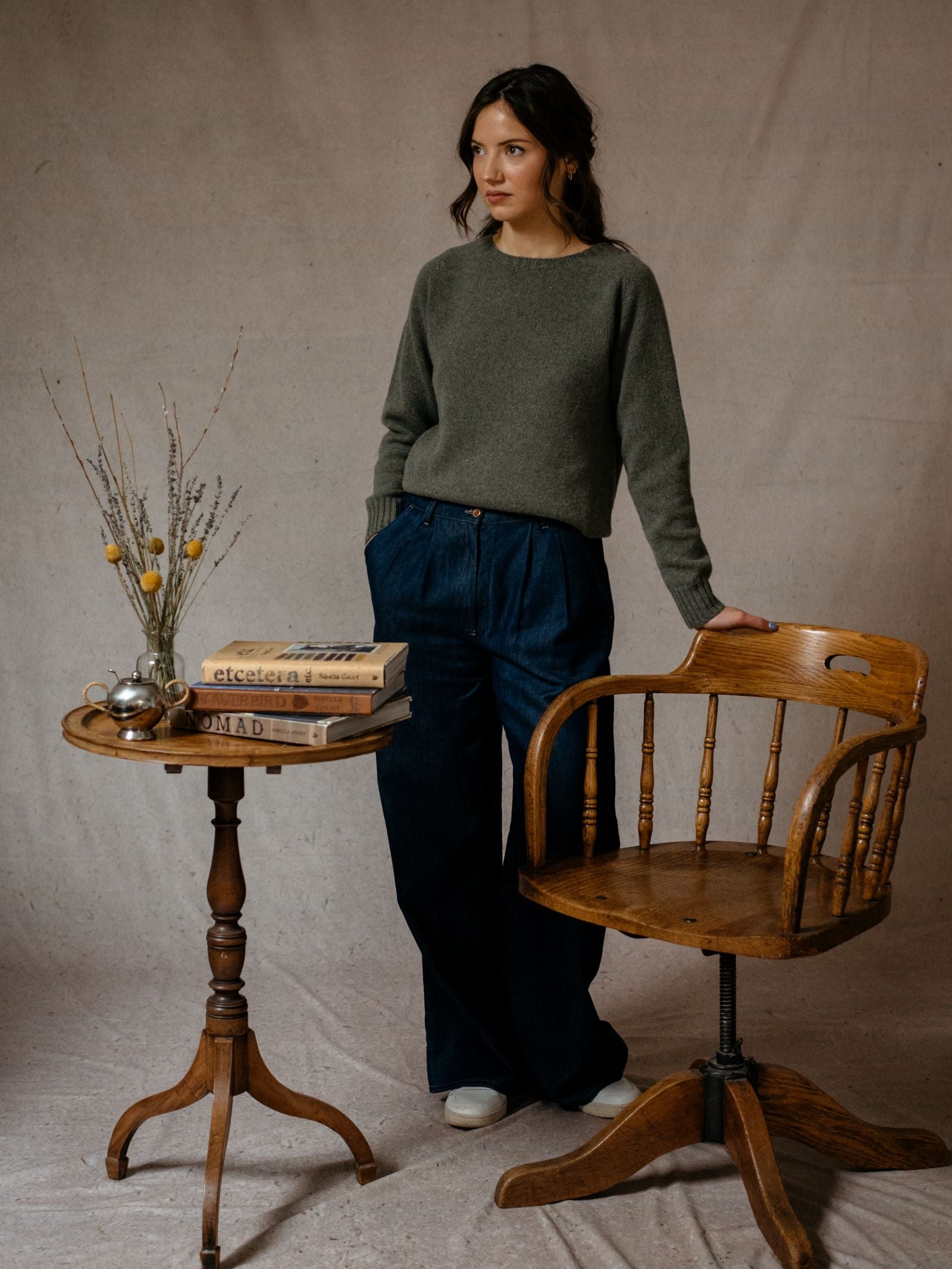 A woman wears the Campbells of Beauly Geelong Lambswool Crew Neck Jumper in gray with wide-leg blue jeans, standing by a wooden chair. Nearby, books, dried flowers, and a teapot rest on a small round table against a beige backdrop.