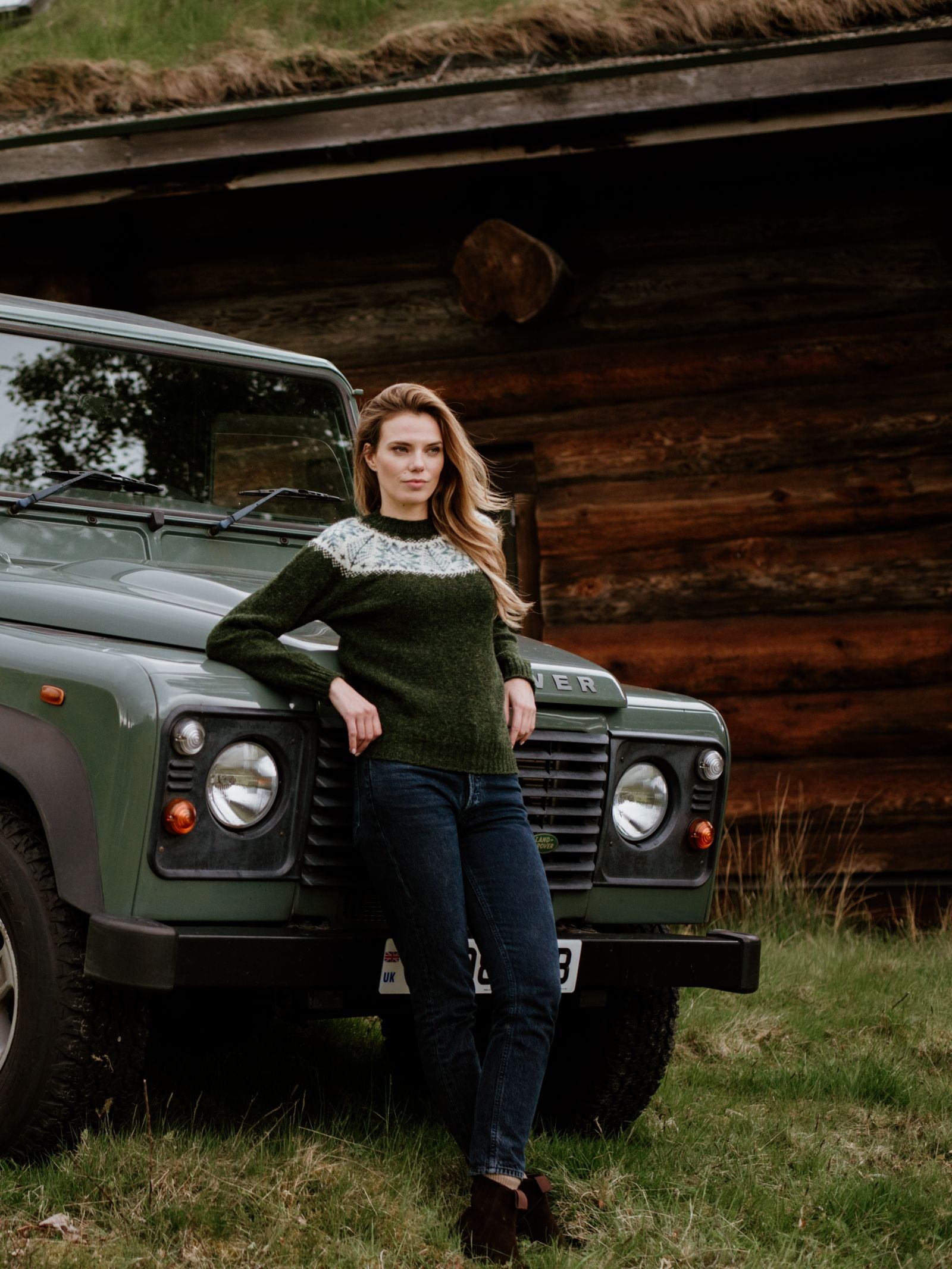 A woman in a green and white Campbells of Beauly Shetland Wool Snowflake Crew Jumper and jeans leans against a green Land Rover on grass, with a rustic wooden building in the background.