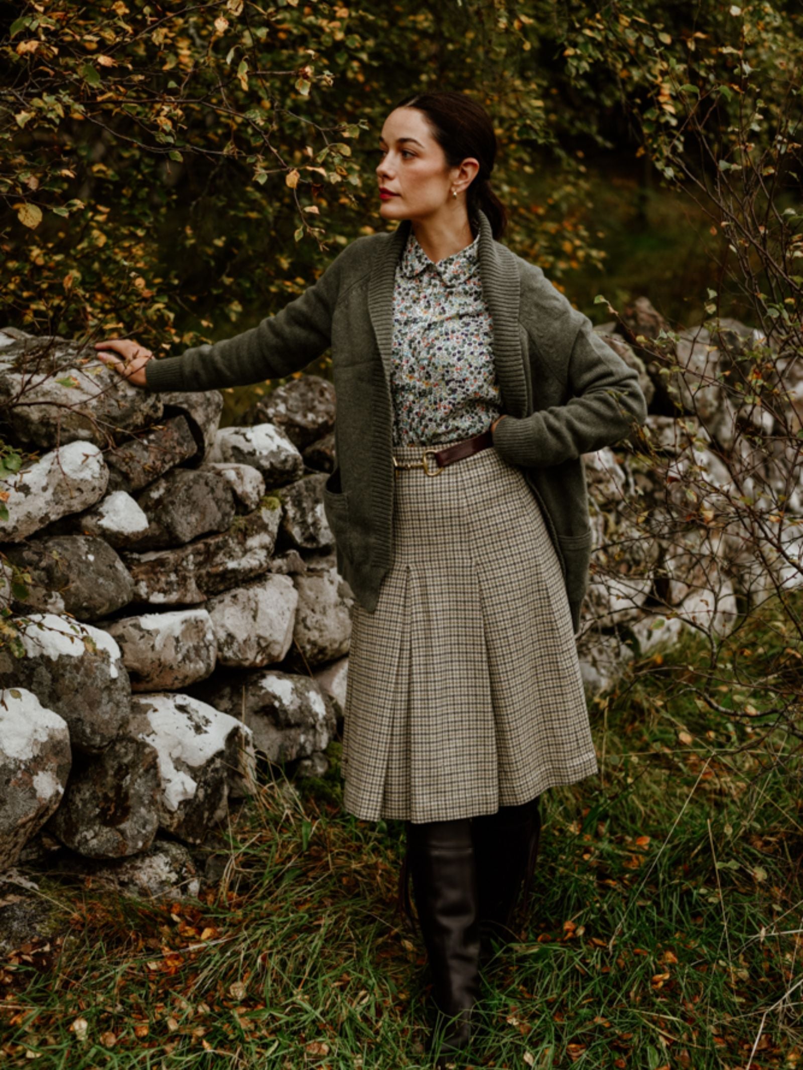 A woman stands outdoors by a stone wall, wearing a floral blouse, plaid skirt, brown belt, black boots, and the Cashmere Shawl Cardigan by Campbell's of Beauly. Autumn leaves and trees surround her.