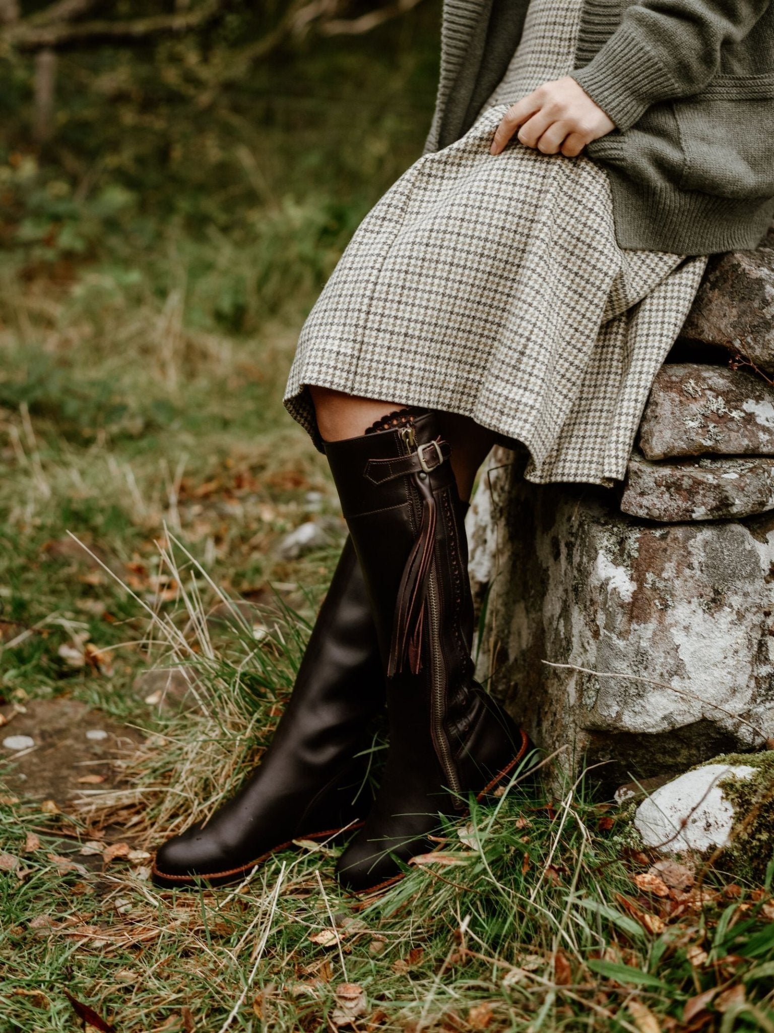 A person wearing Campbell's of Beauly Leather Spanish Boots and a checked dress sits on an outdoor stone wall, surrounded by grass and foliage, with only their lower body visible.