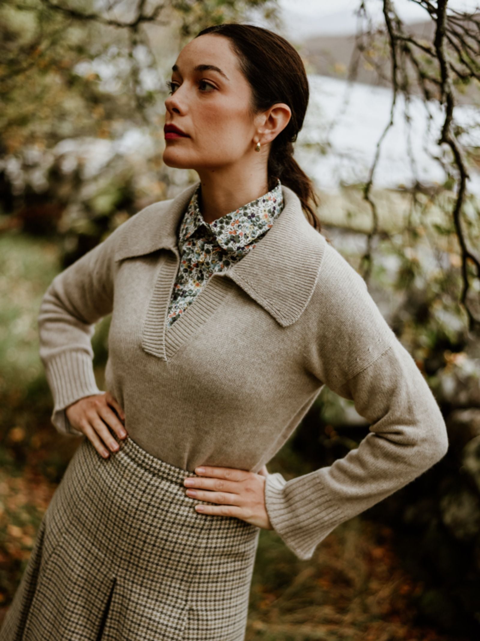 A woman outdoors stands hands-on-hips, wearing the Campbell's of Beauly Cashmere Shirt Jumper layered over a floral blouse and checkered skirt, gazing thoughtfully amid trees and lush greenery inspired by the Scottish Borders.