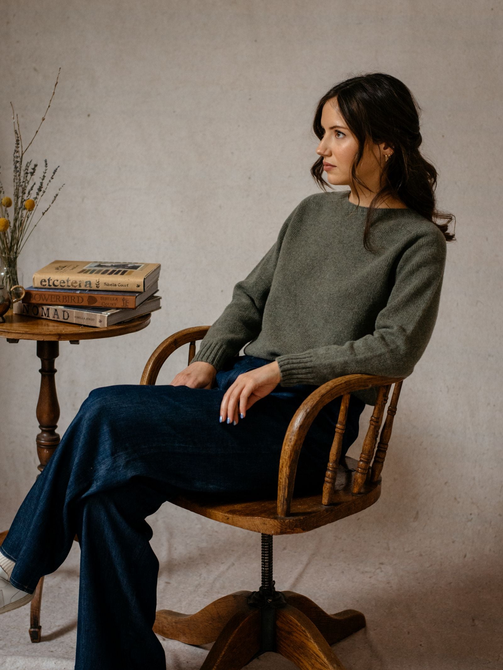 A woman with wavy brown hair sits on a wooden chair, wearing a green Geelong Lambswool Crew Neck Jumper from Campbells of Beauly and dark blue jeans. She gazes to the side. Beside her, a small wooden table holds dried flowers and Scottish books.