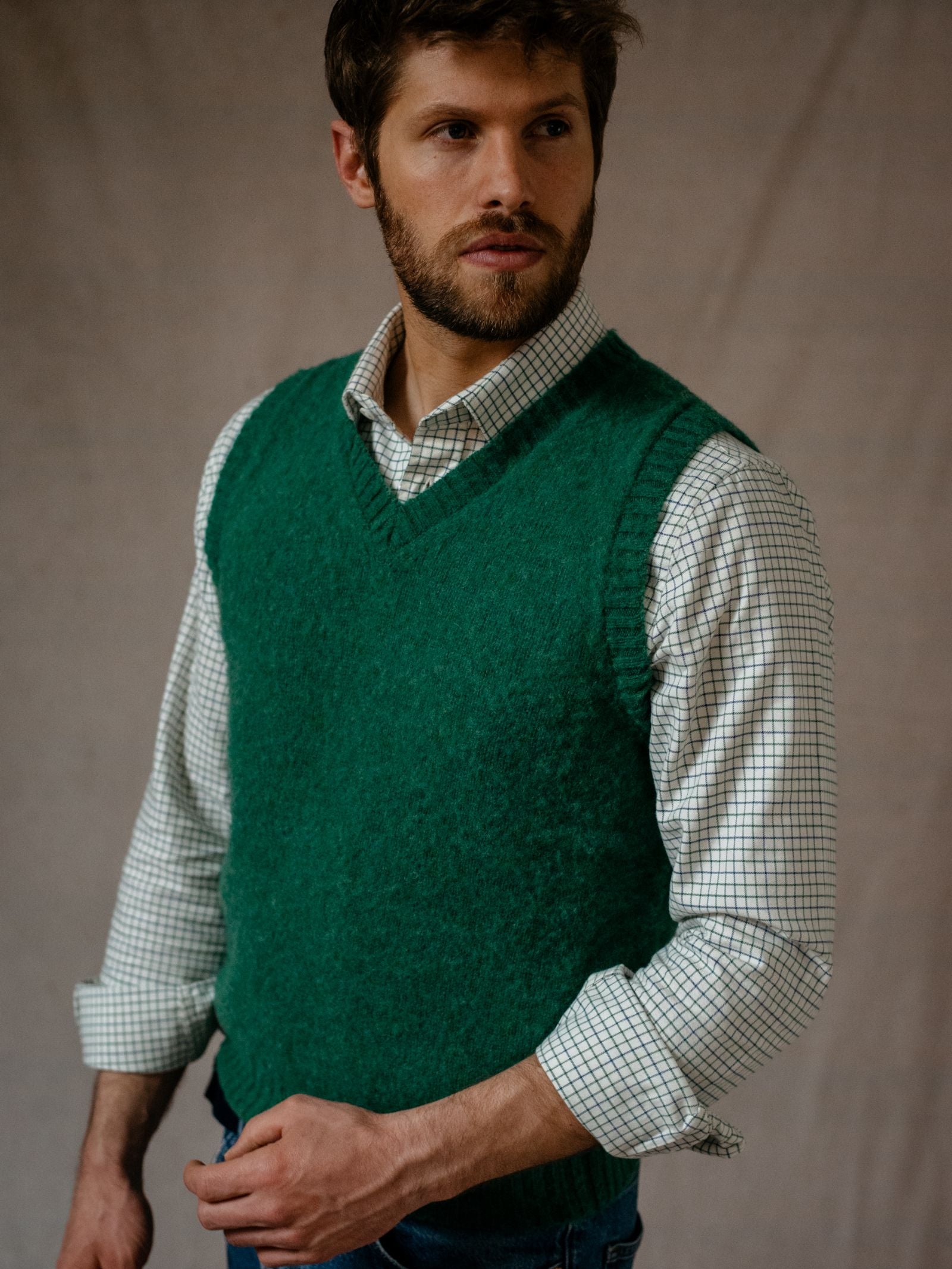 A man with brown hair and a beard wears a green knit sweater vest over a Campbells of Beauly Windowpane Shirt. He stands against a beige background, looking slightly left with a neutral expression and his right hand at his waist.