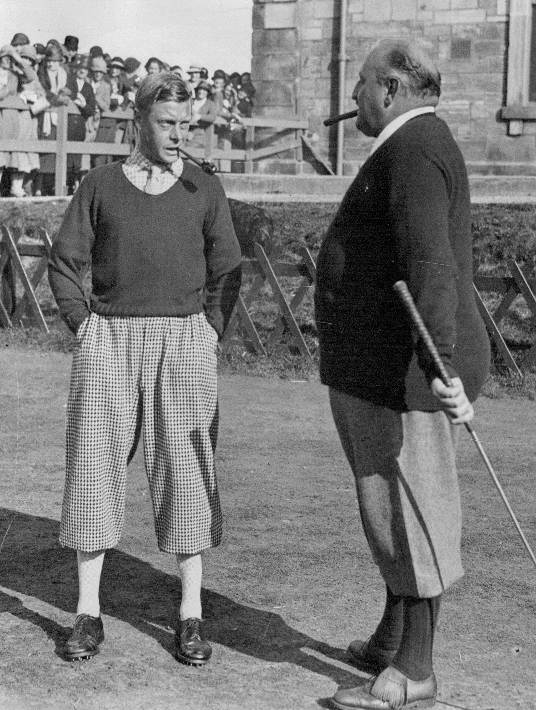 Two men stand outdoors on a golf course in vintage Campbells of Beauly golf attire with knickerbockers. One holds a cigar and club, the other has a pipe. A crowd and stone building can be seen in the background.