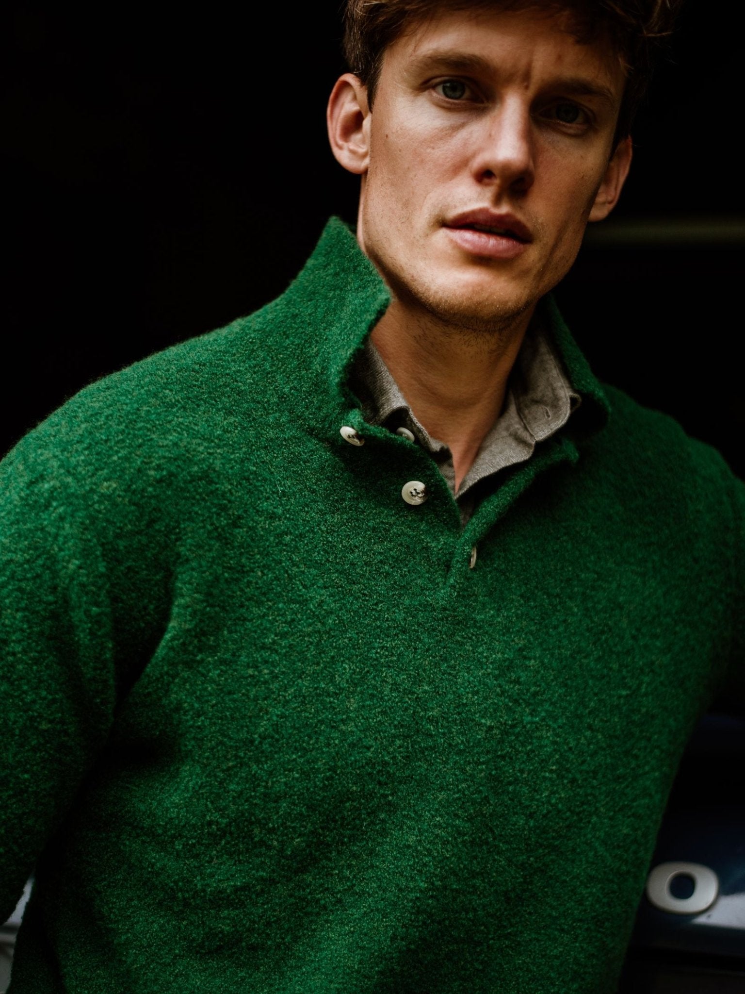 A young man wears the Campbell's of Beauly Bouclé Polo Sweater in textured green over a light gray shirt, standing in soft lighting and gazing past the camera with a neutral expression.