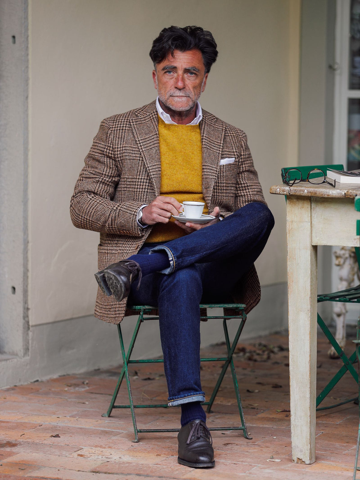A man with dark hair and a beard, wearing the Campbell’s of Beauly Harris Tweed Jacket over a yellow sweater and white shirt, paired with jeans, sits cross-legged on a green chair holding a coffee cup beside a rustic table.