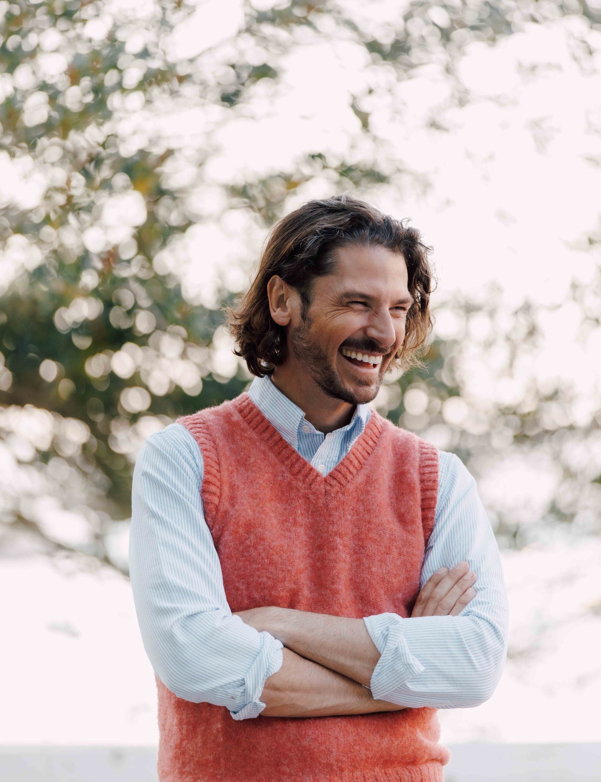 A man with wavy brown hair, wearing a light blue shirt and a sleeveless coral sweater, stands outdoors with arms crossed, smiling and laughing. Blurred trees are in the background.