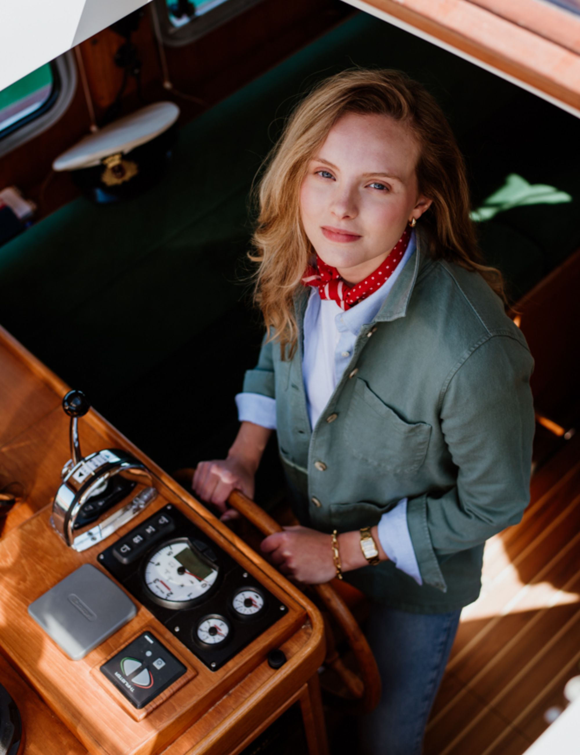 A woman with blonde hair, wearing a green jacket, white shirt, red polka dot scarf, and gold bracelet, stands at the wooden helm of a boat, looking up at the camera. Sunlight shines into the cabin.