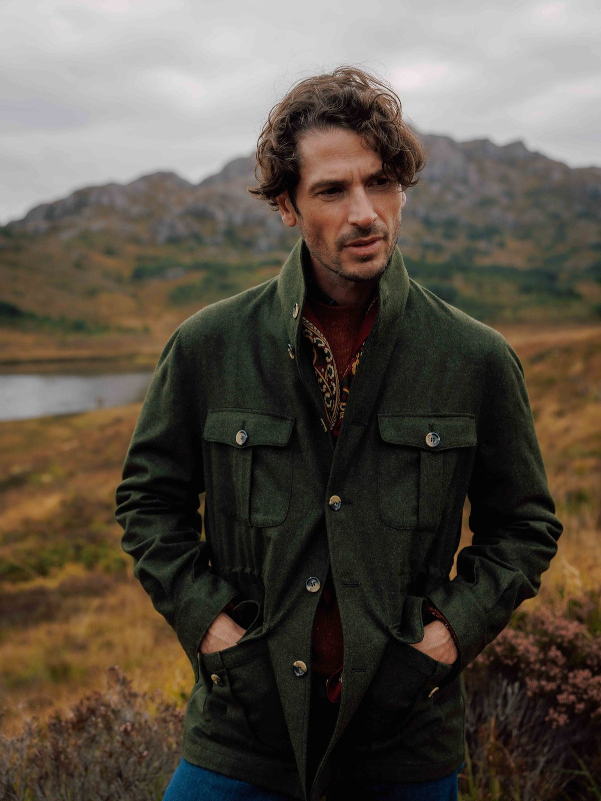 A man wearing a green utility jacket and a patterned jumper stands in a grassy, mountainous landscape.