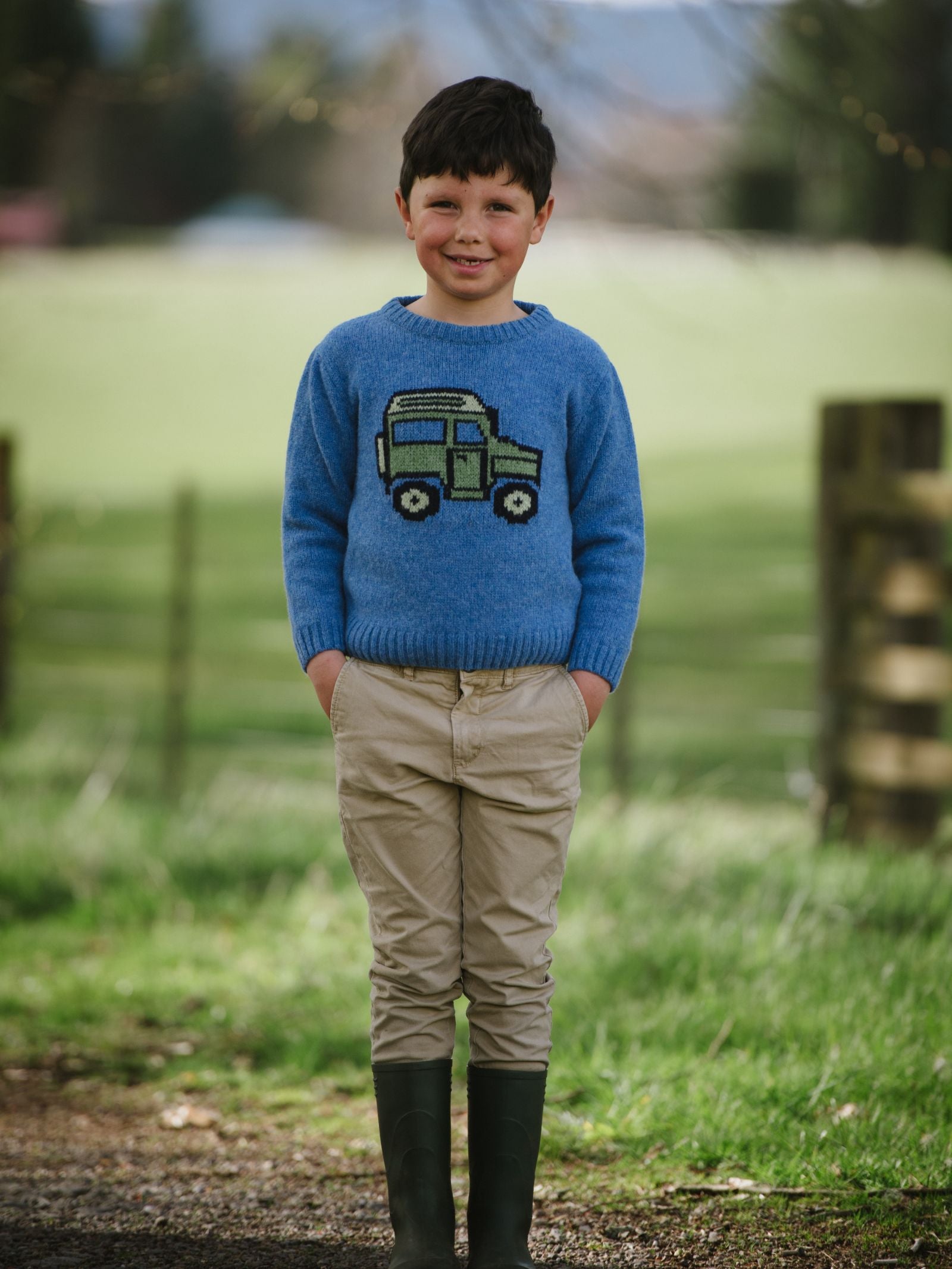 A young boy stands on grass, smiling at the camera in a Campbells of Beauly Mini Land Rover Defender Jumper with a classic green car design, tan pants, and black rain boots. A blurred fence and trees are visible in the background.