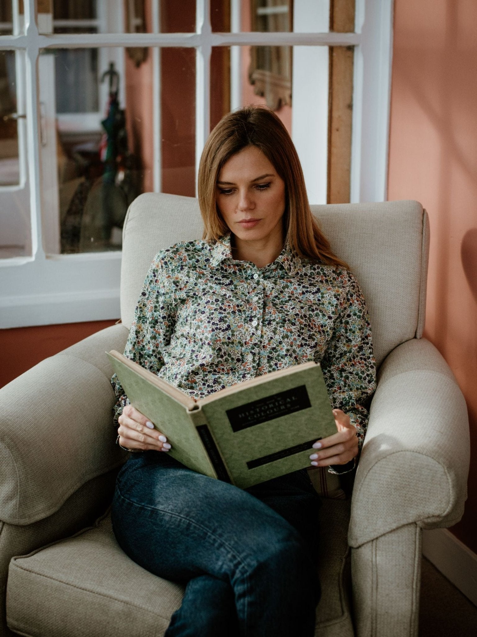 A woman with long hair, dressed in a Campbells of Beauly Liberty Print Cotton Shirt and jeans, sits on a beige armchair reading a green hardcover book in a cozy room with peach walls and white-framed windows.