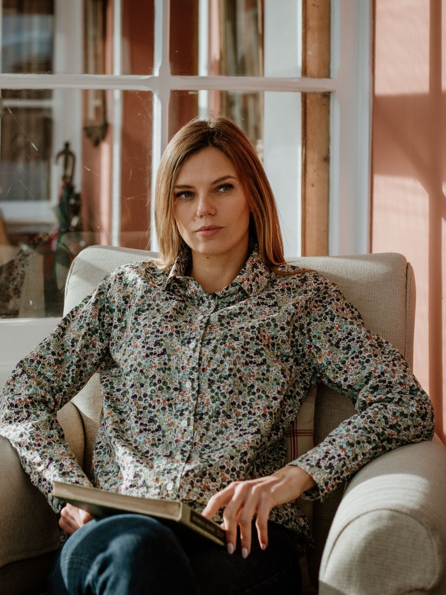 A woman with straight, light brown hair sits by a window in an armchair, wearing a Campbells of Beauly Liberty Print Cotton Shirt, holding an open book and gazing thoughtfully as sunlight streams in.