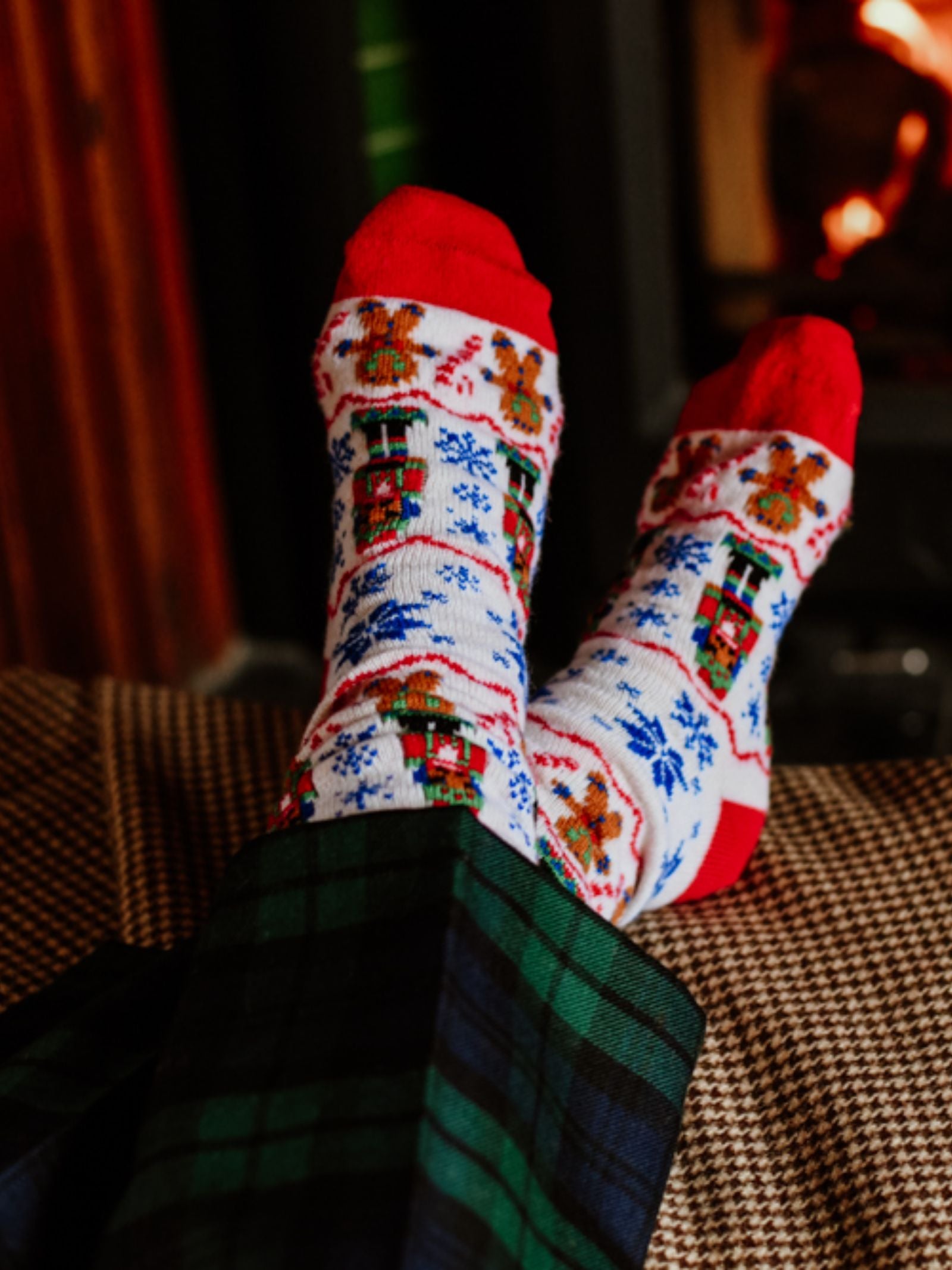 Relaxing by the cozy fireplace, a person showcases Campbell's of Beauly Nutcracker Cashmere Socks—festive footwear with red toes and holiday designs—while wearing plaid pajama pants and putting their feet up.