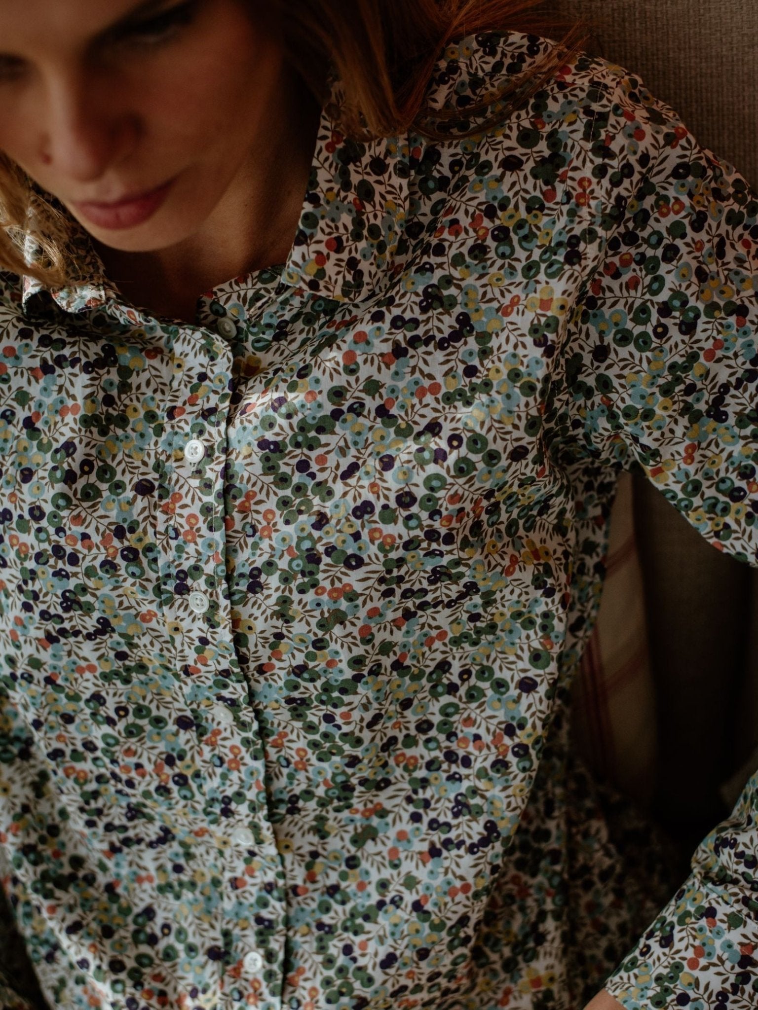 A woman wears the Campbells of Beauly Liberty Print Cotton Shirt, made from vibrant Tana Lawn cotton; soft lighting highlights its exquisite floral pattern as she sits on a chair, partially out of frame.