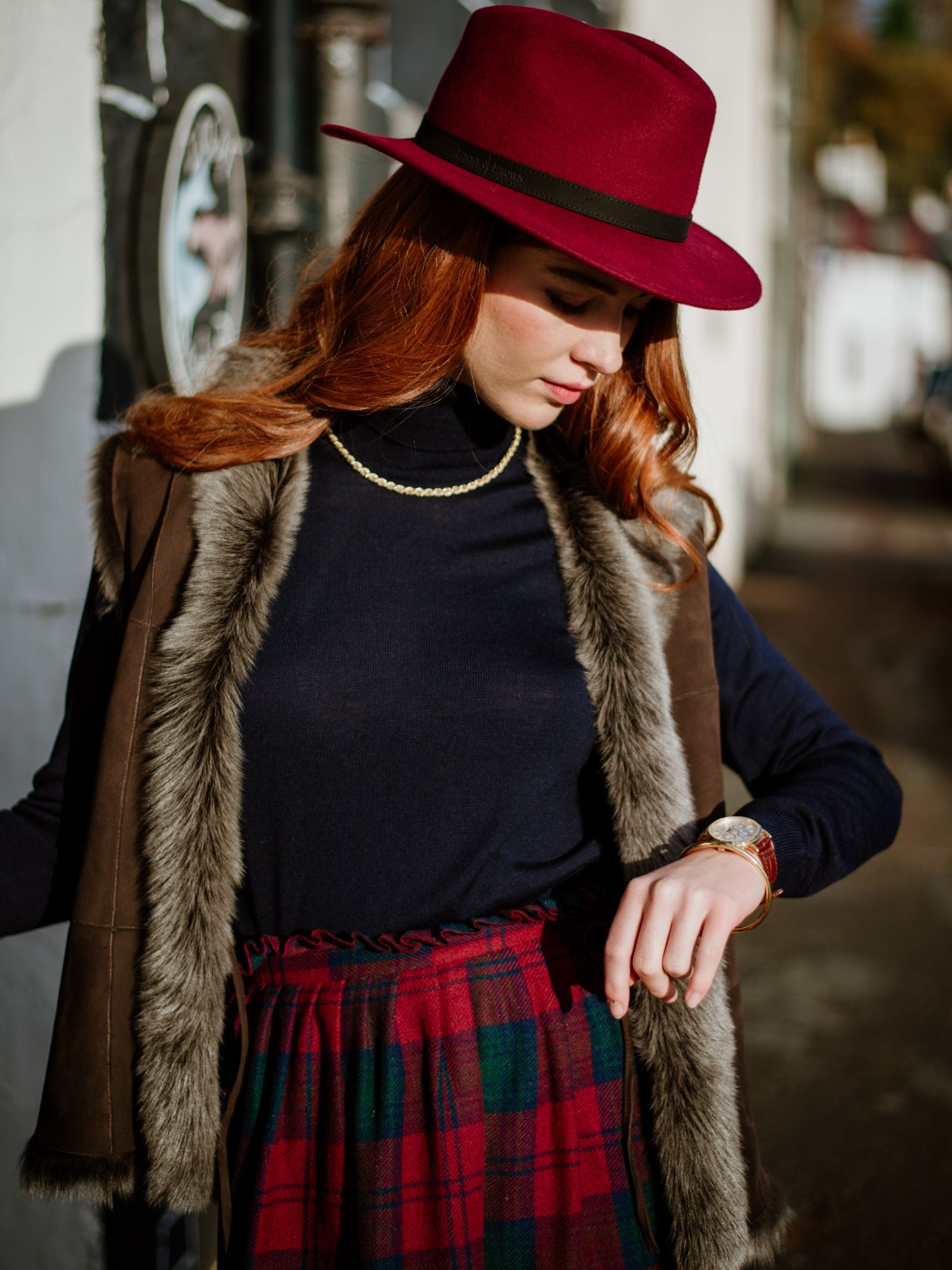 A woman with long red hair wears a Hicks & Brown Gamebird Fedora in burgundy, paired with a black turtleneck, faux fur vest, plaid skirt, and gold necklace. Outdoors on a sunny day, she glances down at her watch.