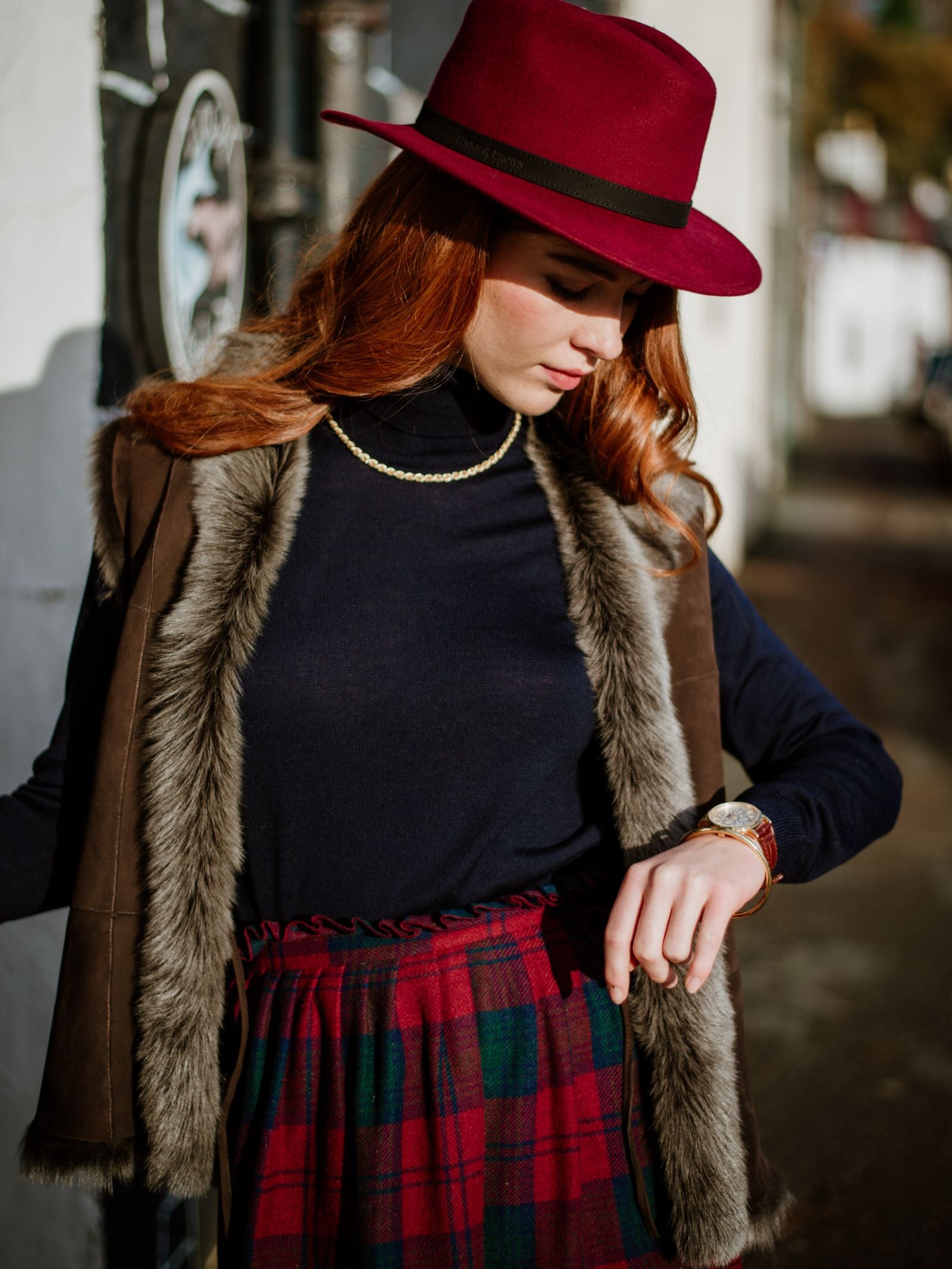 A woman with long red hair wears a red wide-brimmed hat, a Campbell’s of Beauly Sheepskin Reversible Gilet over a navy turtleneck, a gold necklace, and a red and green plaid skirt. She stands outdoors, looking down at her watch.