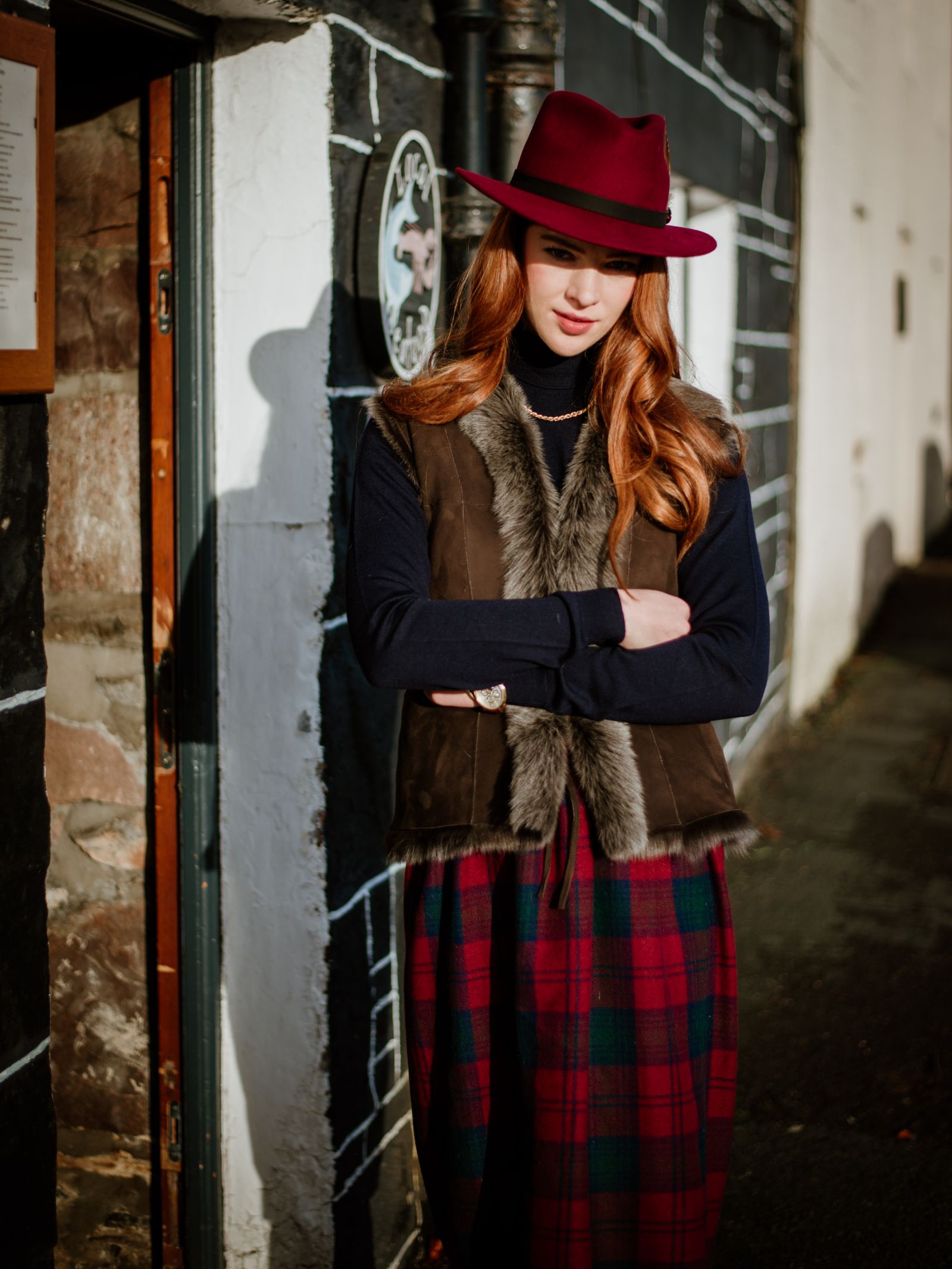 A woman with long red hair, wearing a burgundy hat and a Campbells of Beauly Sheepskin Reversible Gilet, stands outside against a stone and white wall in sunlight, arms crossed over her red and green plaid skirt.