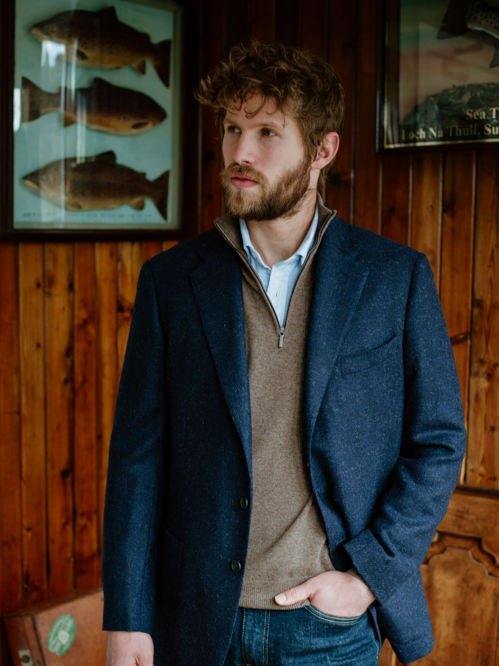 A bearded man with curly hair stands indoors, wearing the Campbells of Beauly Navy Twill House Jacket over a brown sweater and blue shirt. Wooden walls and framed fish illustrations are behind him as he looks to the side with one hand in his pocket.