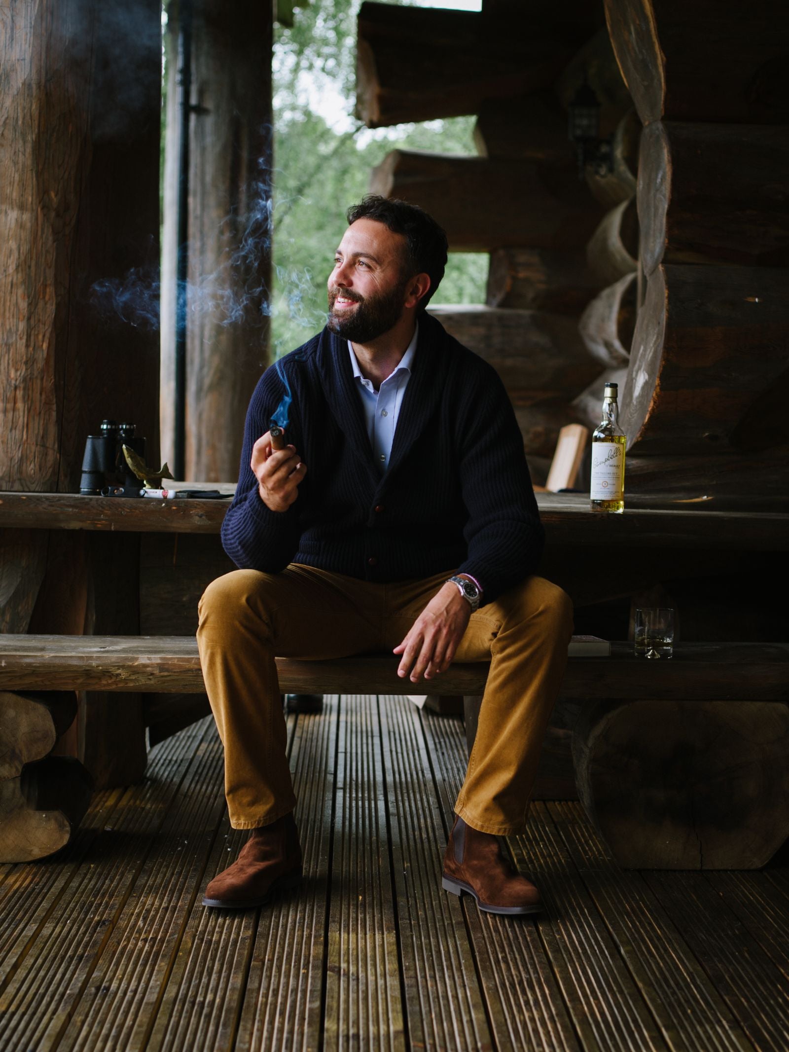 A bearded man in the Campbells of Beauly Shawl Collar Cardigan and brown pants sits on a wooden porch, smiling with a smoking pipe. Nearby are a bottle, glass, and binoculars; logs and greenery complete this cozy winter scene.