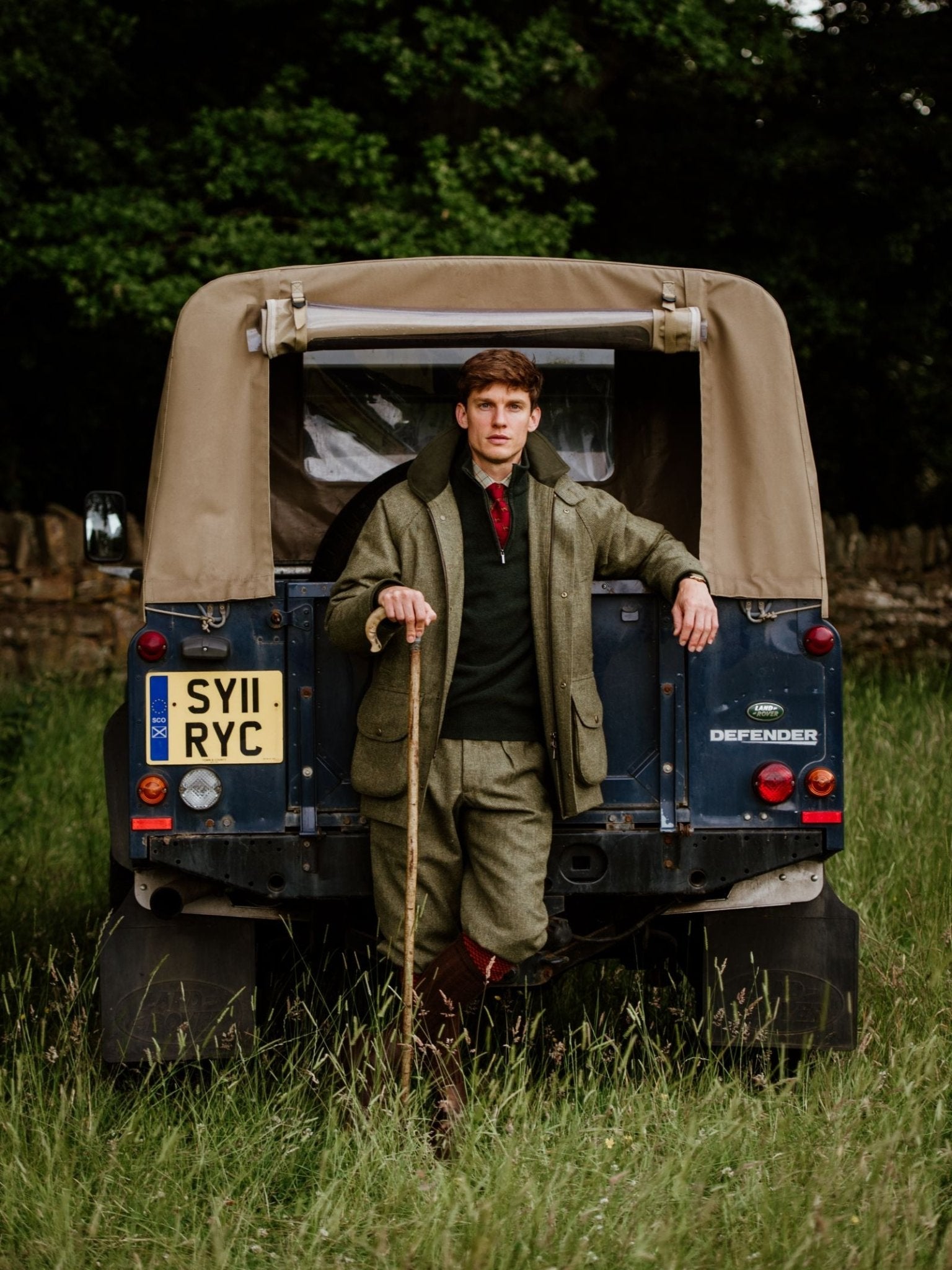A man in country attire and Campbells of Beauly Tweed Breeks leans against a Land Rover Defender parked in tall grass, holding a wooden walking stick, with trees and a stone wall in the background.
