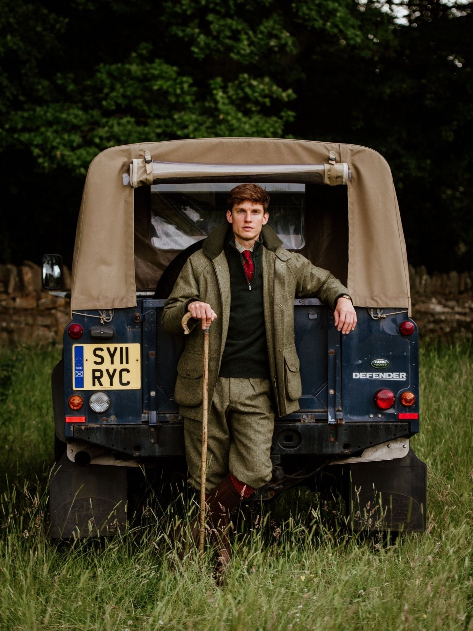 A man in country attire and a Campbells of Beauly Tweed Shooting Waistcoat leans on a walking stick beside a vintage Land Rover Defender in a grassy field with trees and a stone wall—ideal for countryside pursuits.
