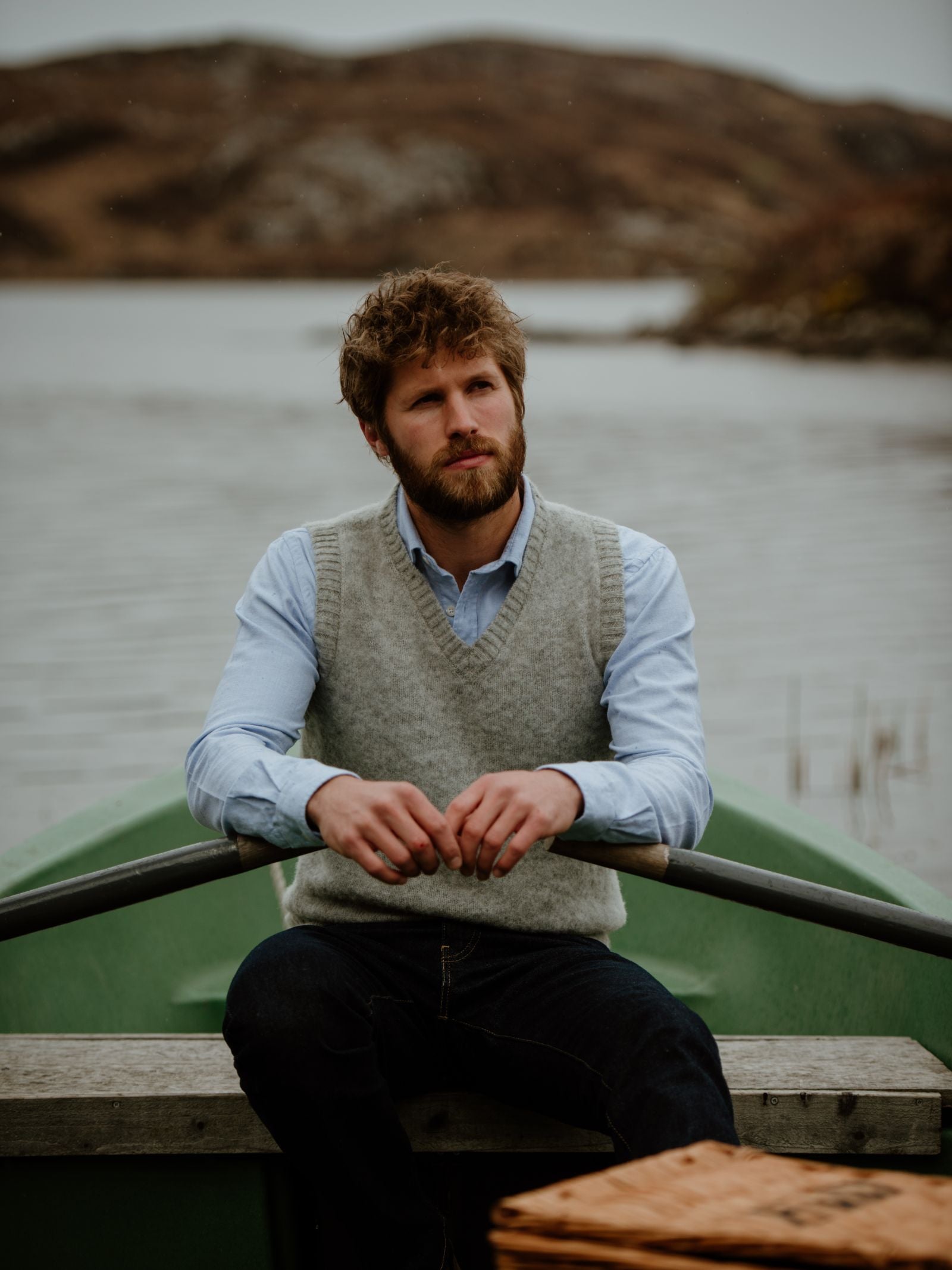 A man with a beard and curly hair sits on a green rowboat by a lake, wearing the Campbells of Beauly Shetland Slipover layered over a gray vest, with misty hills and water in the background on a cloudy day.