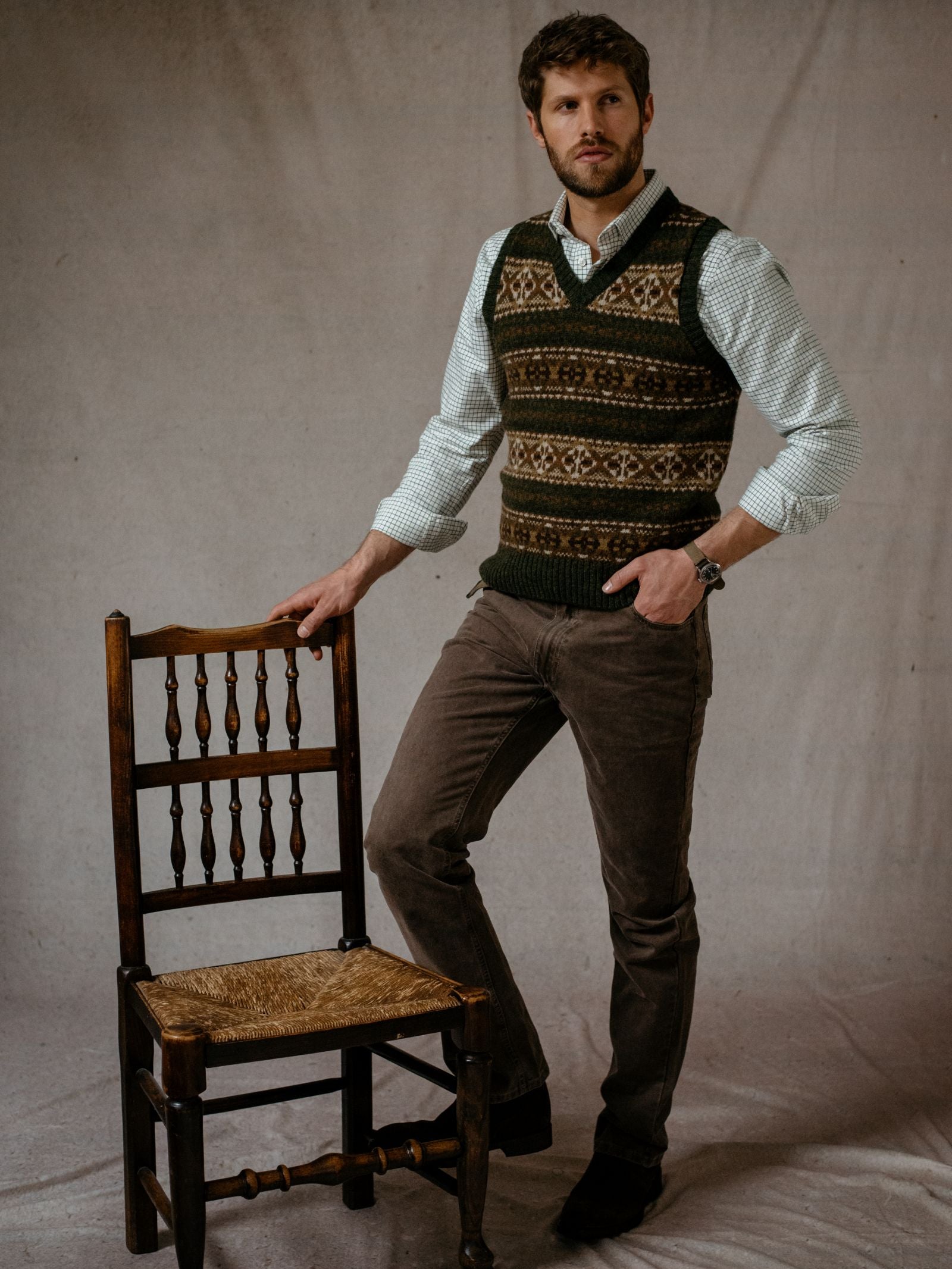 A man with short brown hair and a beard stands beside a wooden chair, wearing the Fairisle Slipover by Campbells of Beauly over a light shirt and brown pants. He rests one hand on the chair against a plain, neutral backdrop.