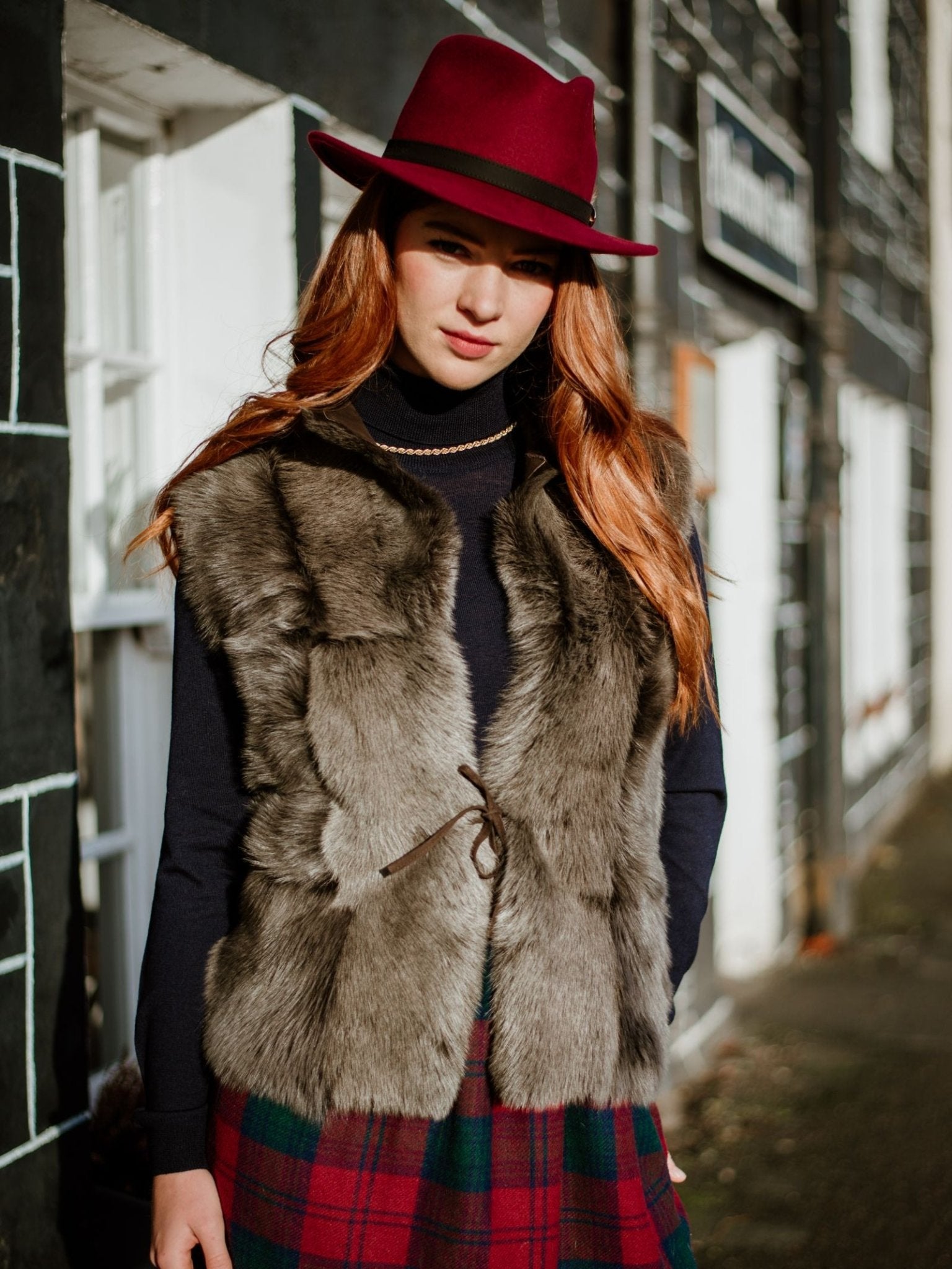 A woman with long red hair stands outdoors by a stone building in sunlight, wearing a burgundy hat, a Campbells of Beauly Sheepskin Reversible Gilet over a black turtleneck, and a red-and-green plaid skirt.